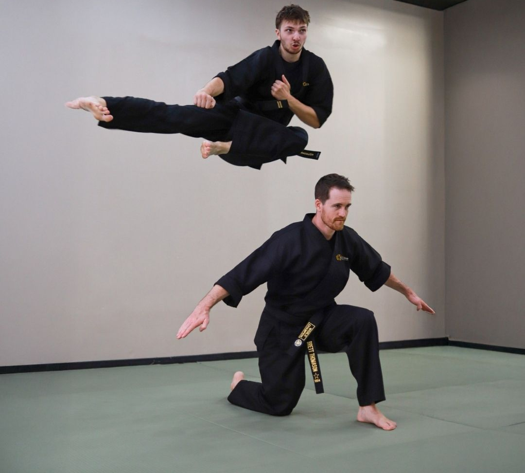 Children in colorful gis play a game on a mat. Two instructors pull a student in blue.