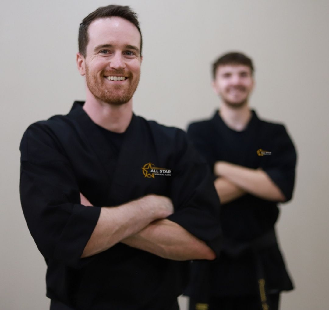 People practicing Brazilian Jiu-Jitsu. One person on the ground, another on top; two others grappling nearby. Black gis, indoor setting.
