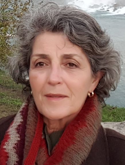 Mujer con cabello gris y rizado, ojos marrones y una bufanda estampada, al aire libre junto al agua.