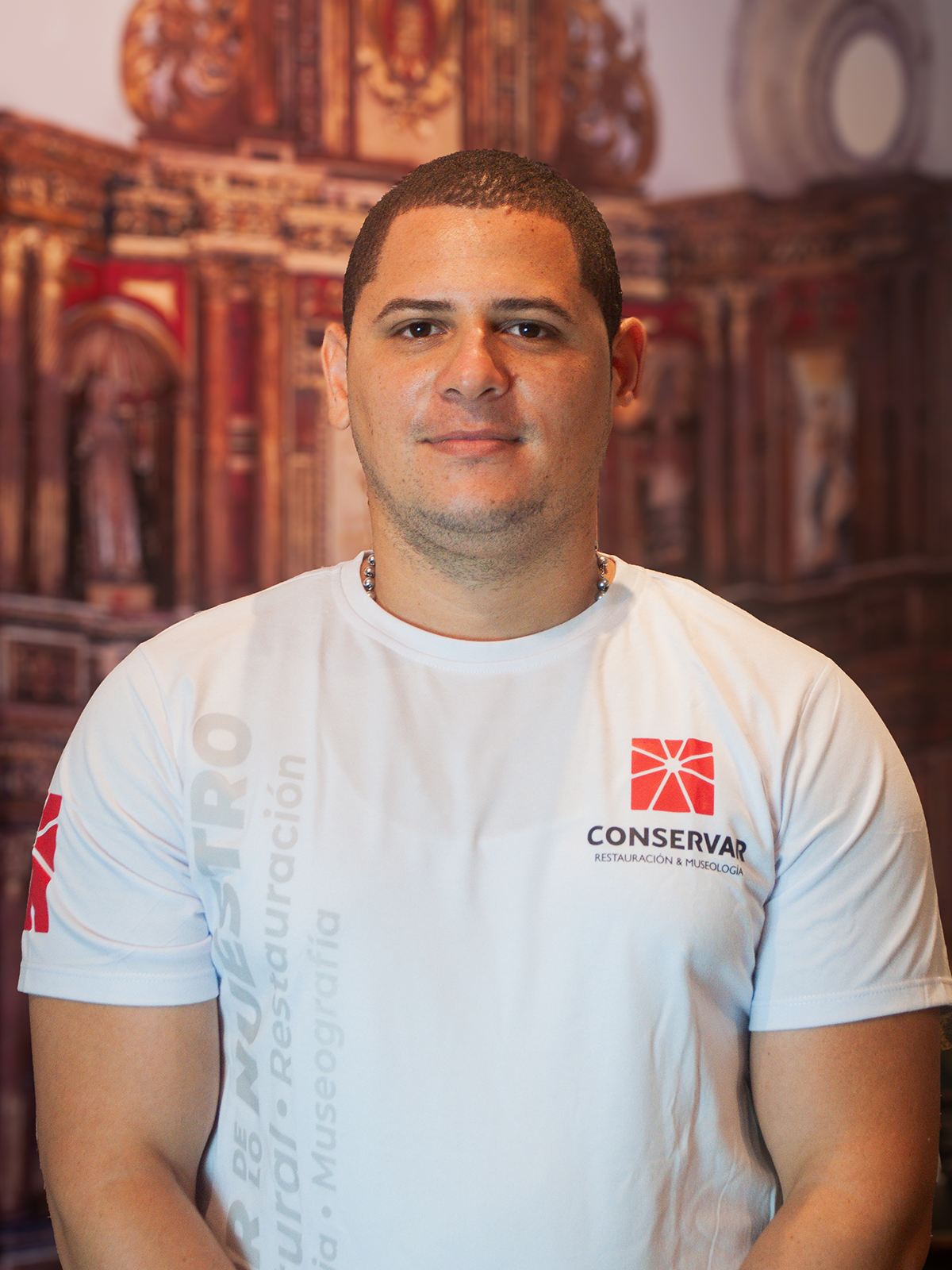 Hombre con camiseta blanca con logo, sonriendo, de pie frente a un fondo arquitectónico borroso.