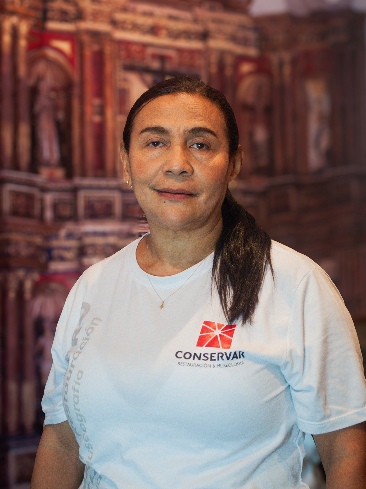 Mujer con camiseta blanca y coleta, con el logo de CONSERVVI. Sonriendo, frente a un fondo arquitectónico borroso.