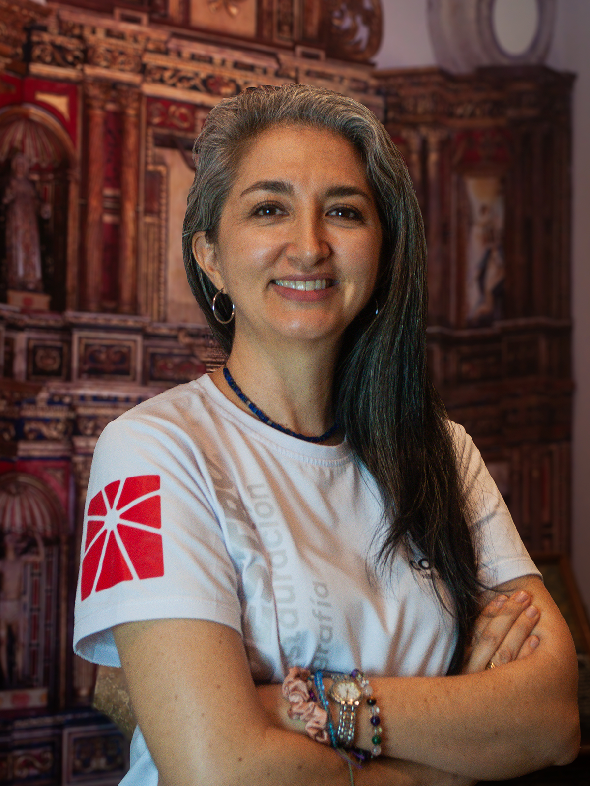 Mujer de cabello gris y brazos cruzados sonriendo, vistiendo una camisa blanca con un logo rojo, frente a un fondo de madera.