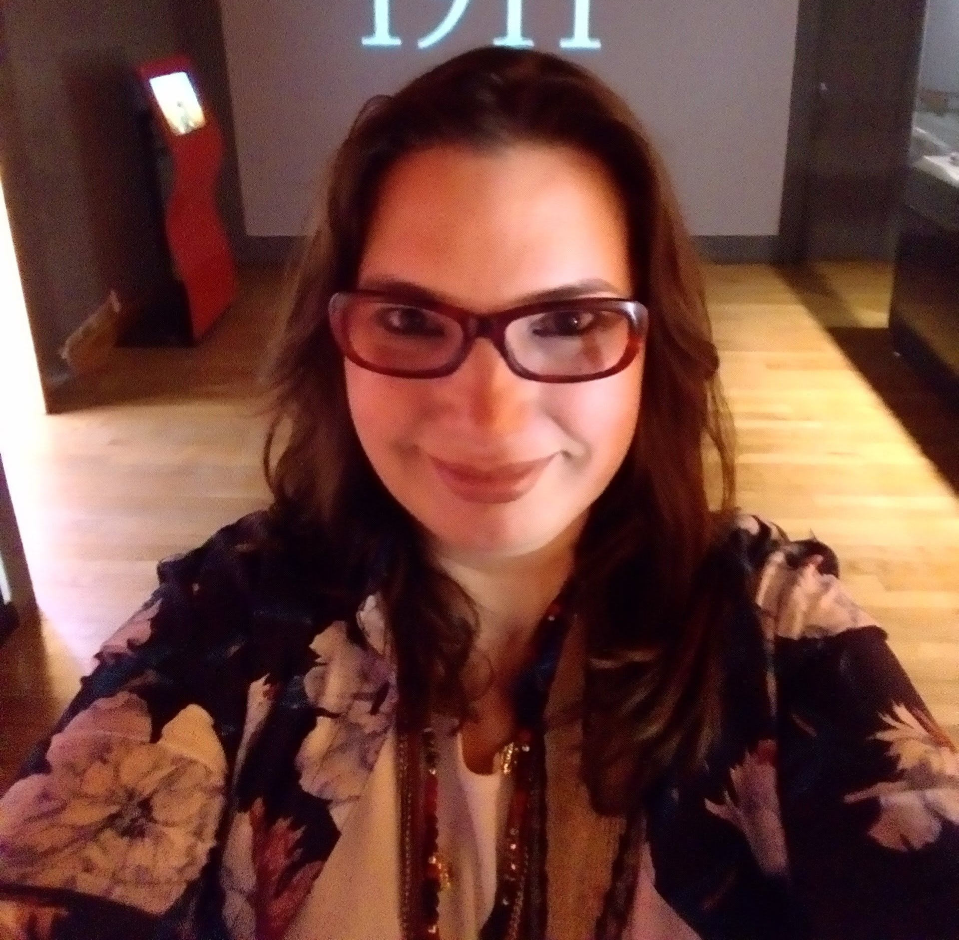 Mujer con gafas sonríe a la cámara, vistiendo una chaqueta con estampado floral, en un museo.