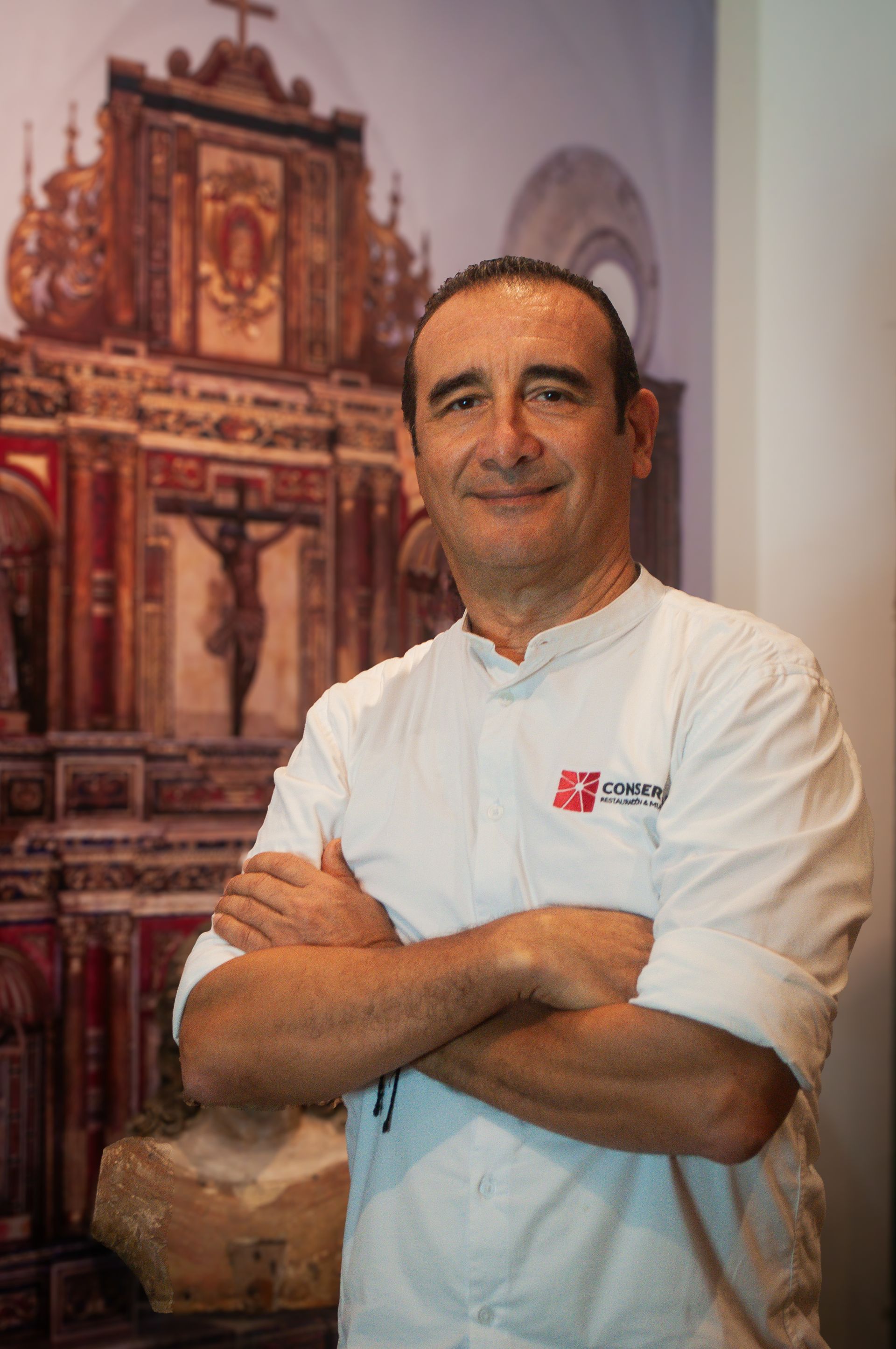 Un hombre con chaqueta blanca de cocinero, con los brazos cruzados, sonríe delante de un cuadro de una iglesia.