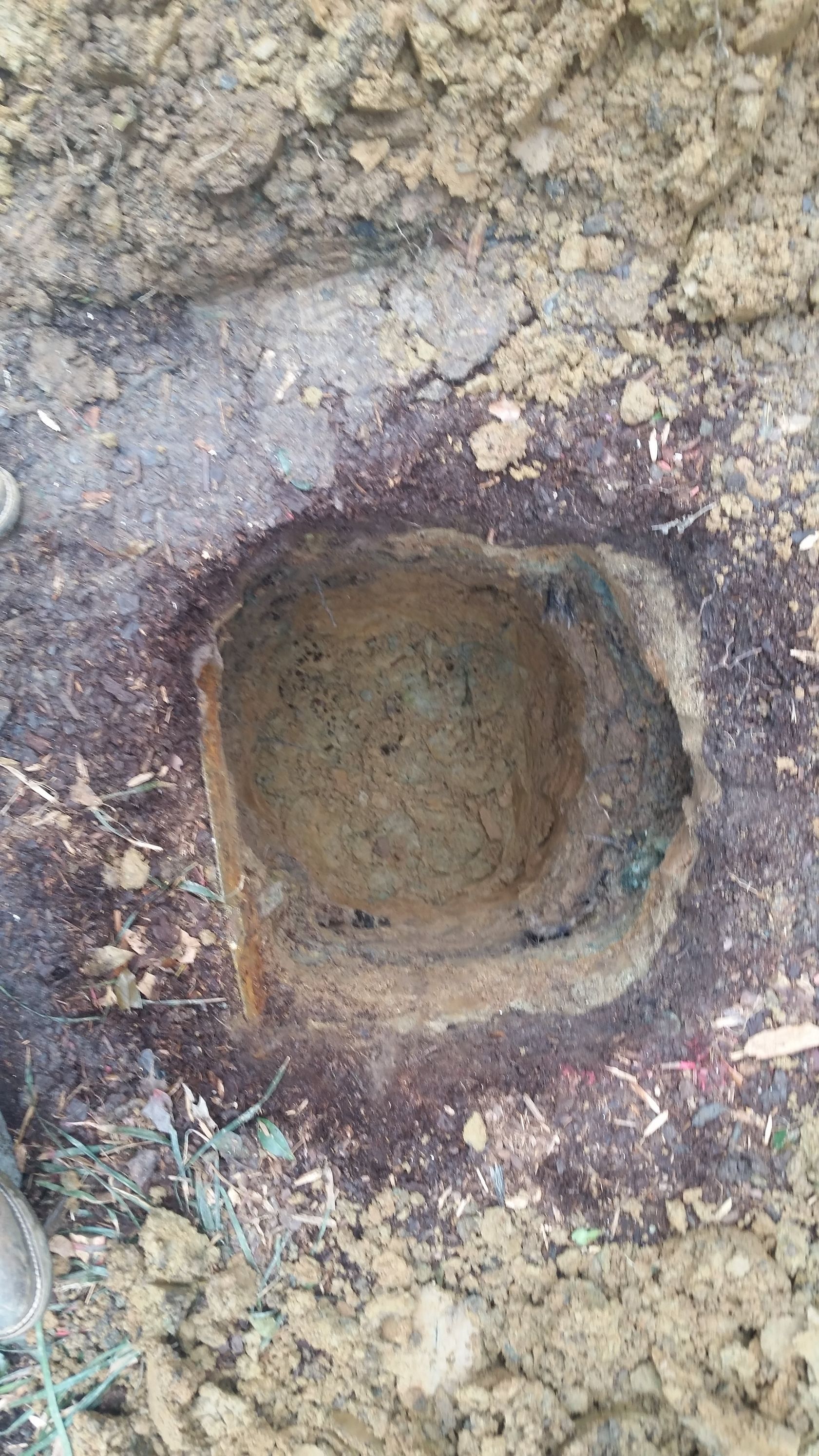 Square hole dug in dark soil, possibly for planting, surrounded by reddish-brown stains and wood chips.