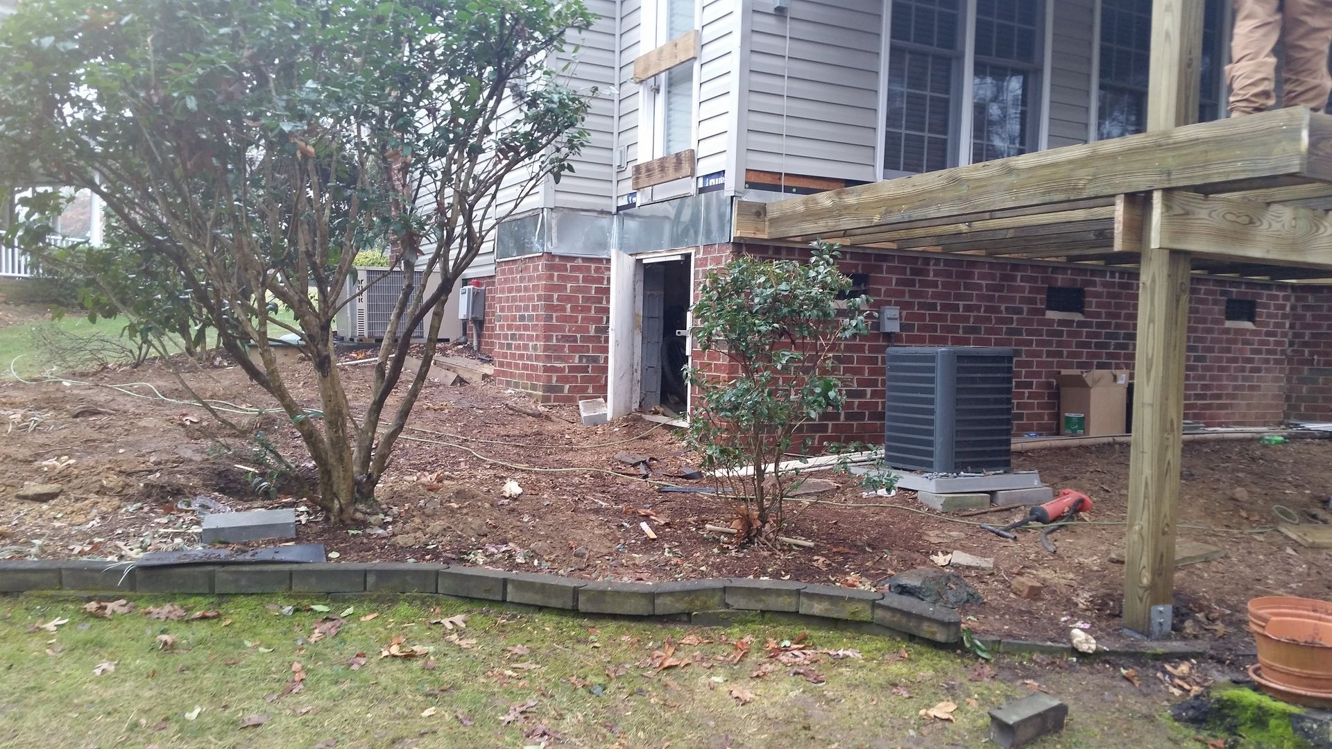 A building with brick and white siding, a deck, and yard with shrubs, landscaping, and a dark AC unit.
