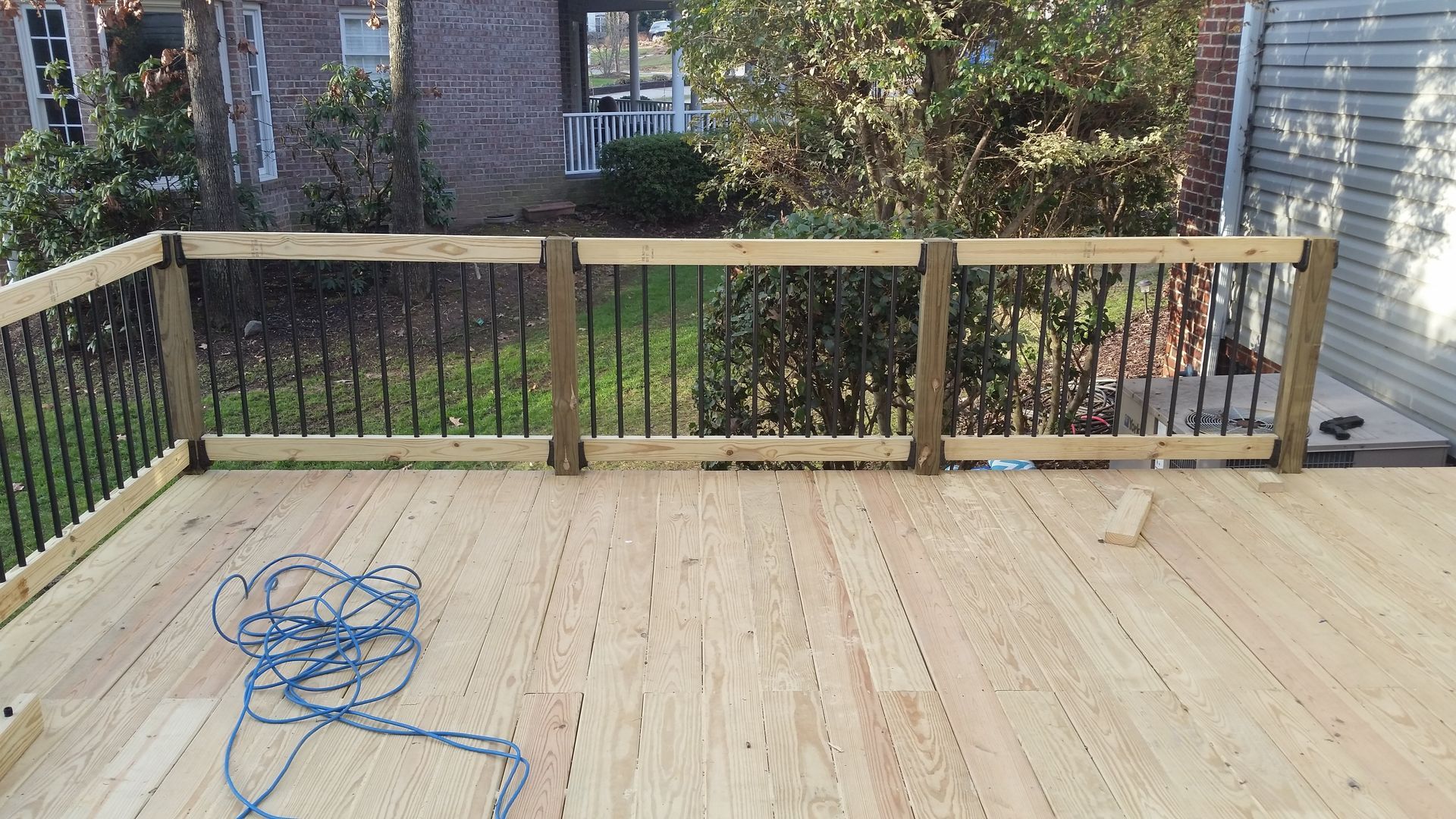 Wooden deck with black railing and blue rope in foreground; brick house and trees in background.