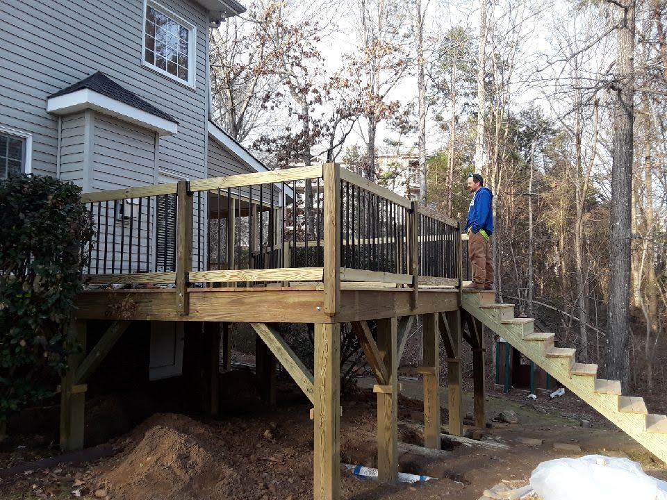 Wooden deck with stairs, built on posts. Person stands on deck, looking outward. Trees in background.
