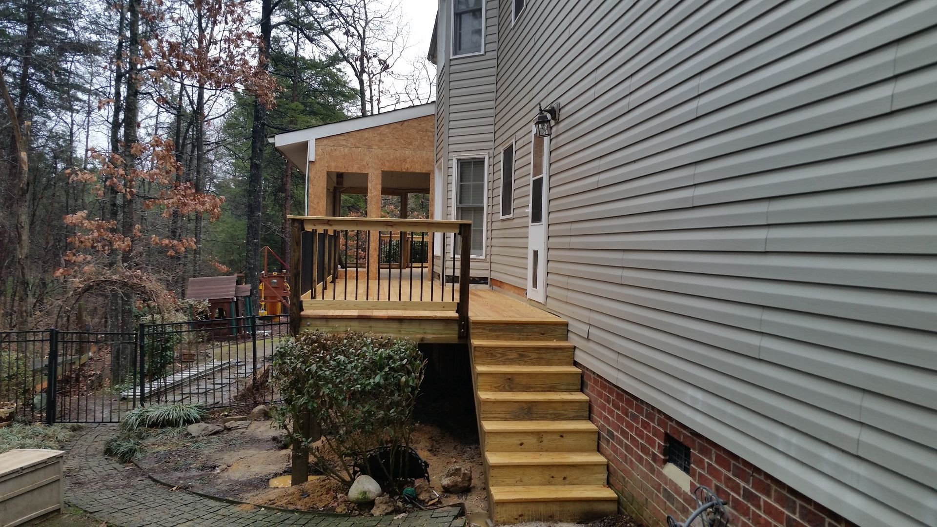 Back of a house with a wooden deck and stairs; unfinished structure visible, surrounded by trees.