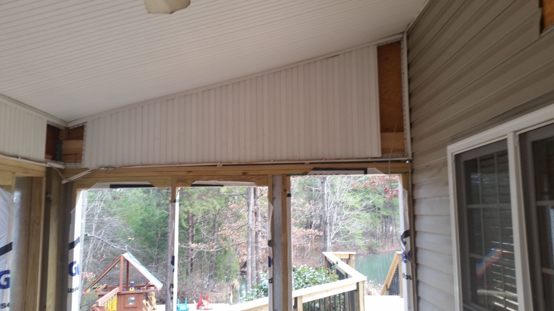 Sunroom interior under construction, white panel ceiling and partial wall. Sliding glass doors lead to deck overlooking trees.