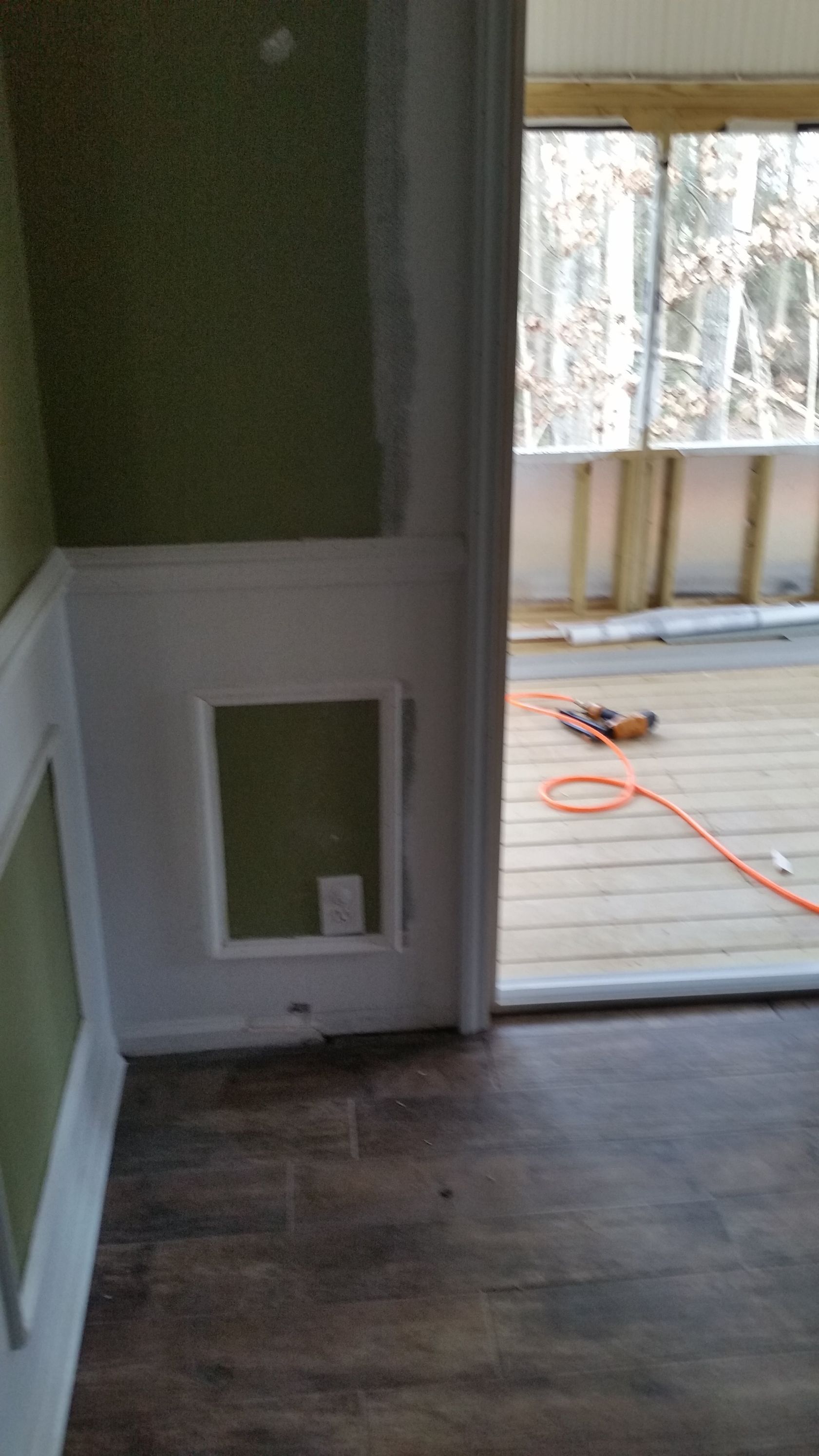 Interior doorway with wainscoting and a cut-out square, leading to a wood-planked deck with construction tools.