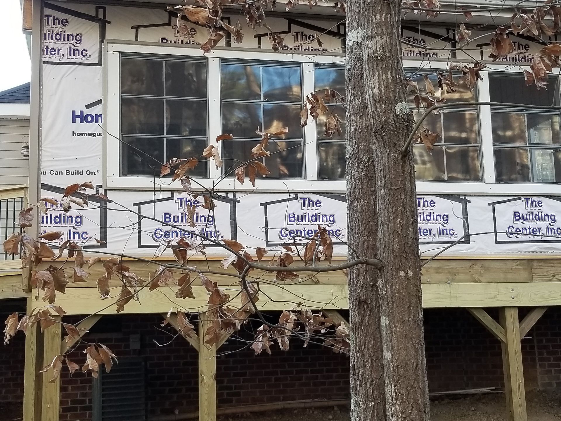 New construction: windows on a house with a wooden deck, partially obscured by a tree with brown leaves.