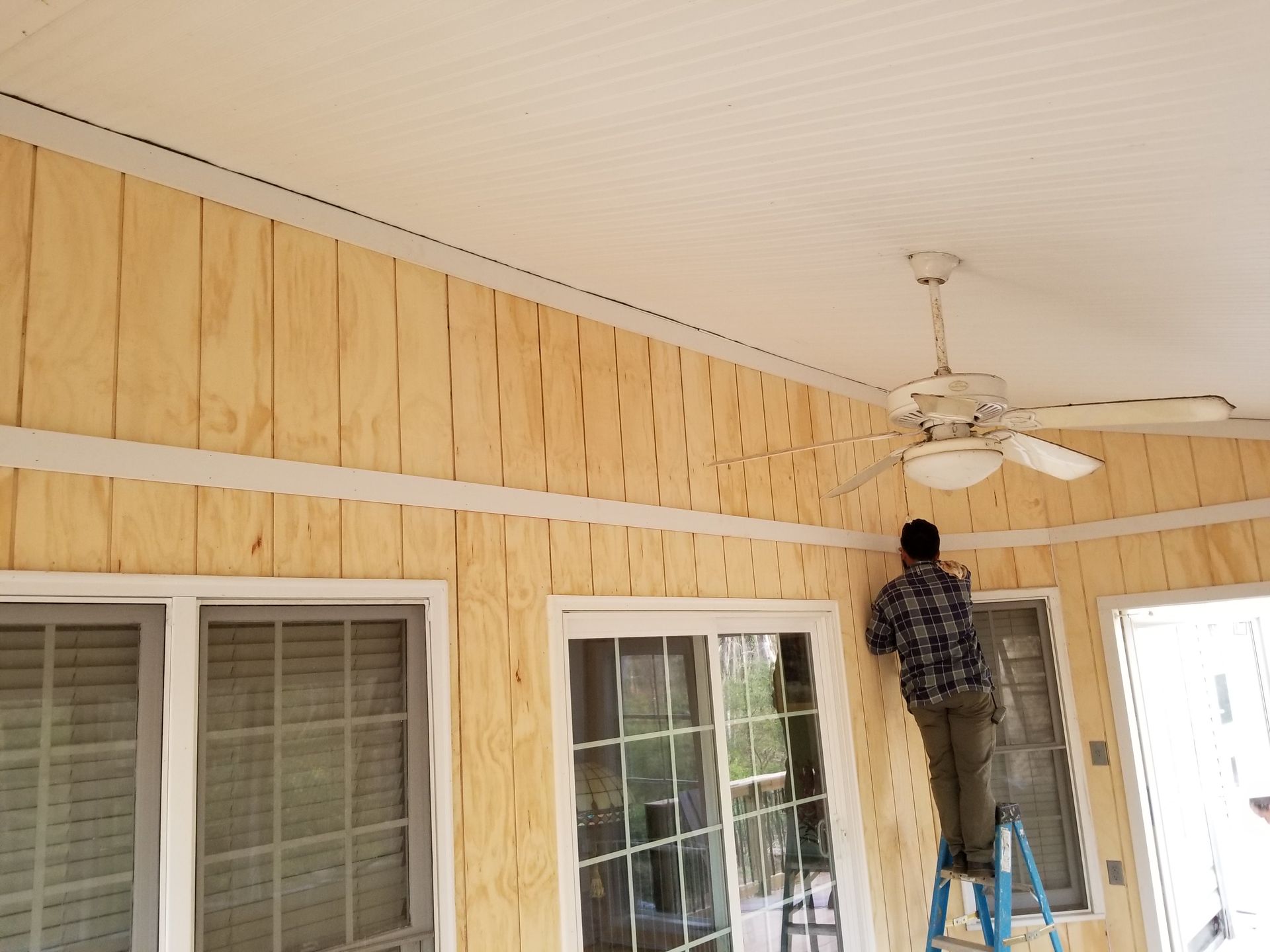 Person on a ladder installs trim on a wood-paneled wall, outdoors under a white ceiling with a fan.