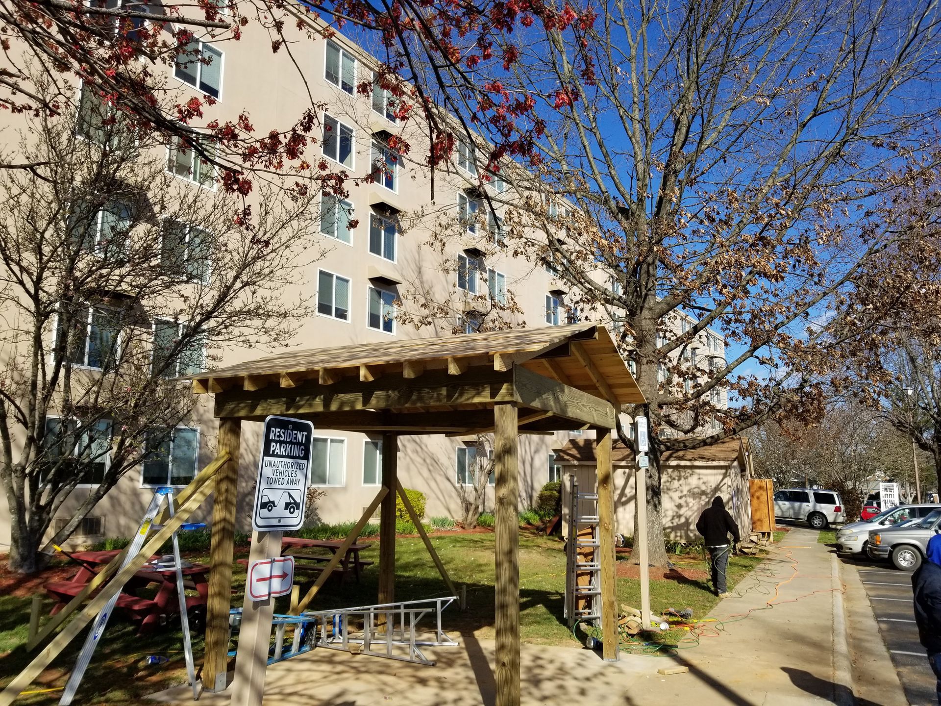 Wooden structure with a roof, next to a building. Sidewalk and parked cars. Trees with bare branches.