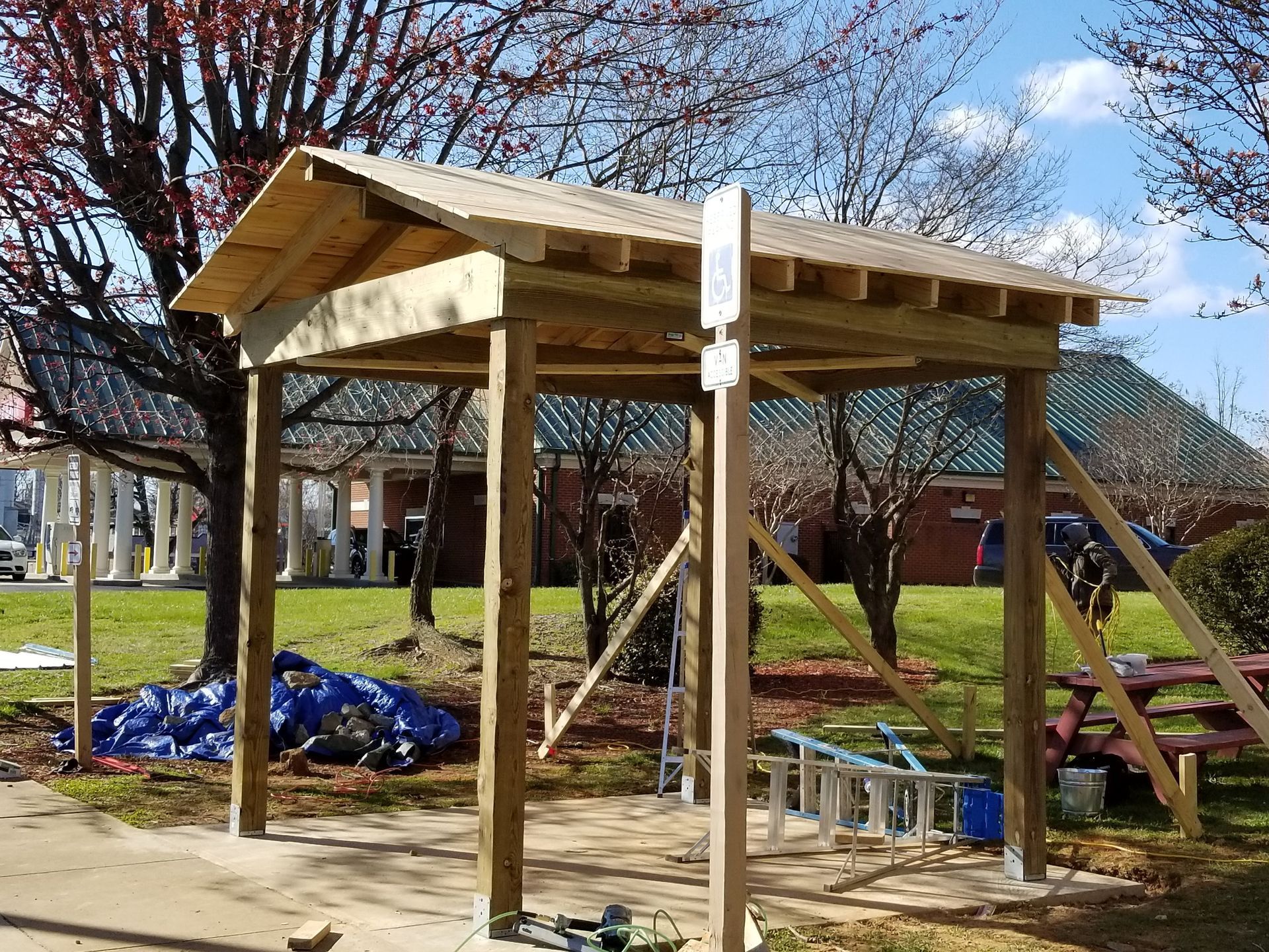 Wooden shelter being built outdoors with a pitched roof, supporting posts, and construction materials nearby.