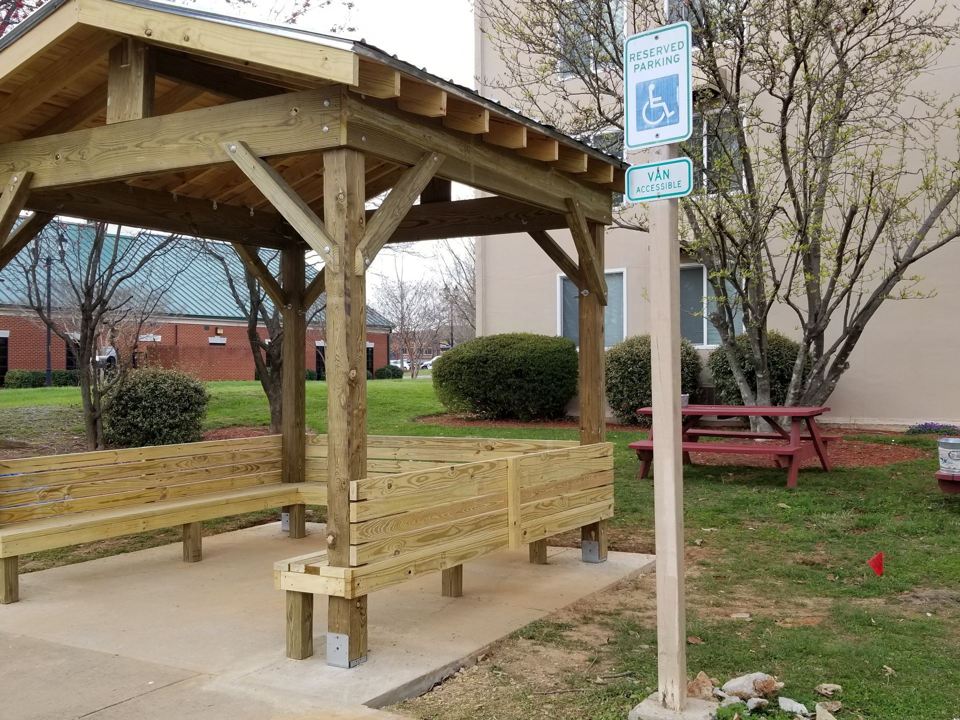 Wooden gazebo with benches, accessible parking sign, and a building in the background.