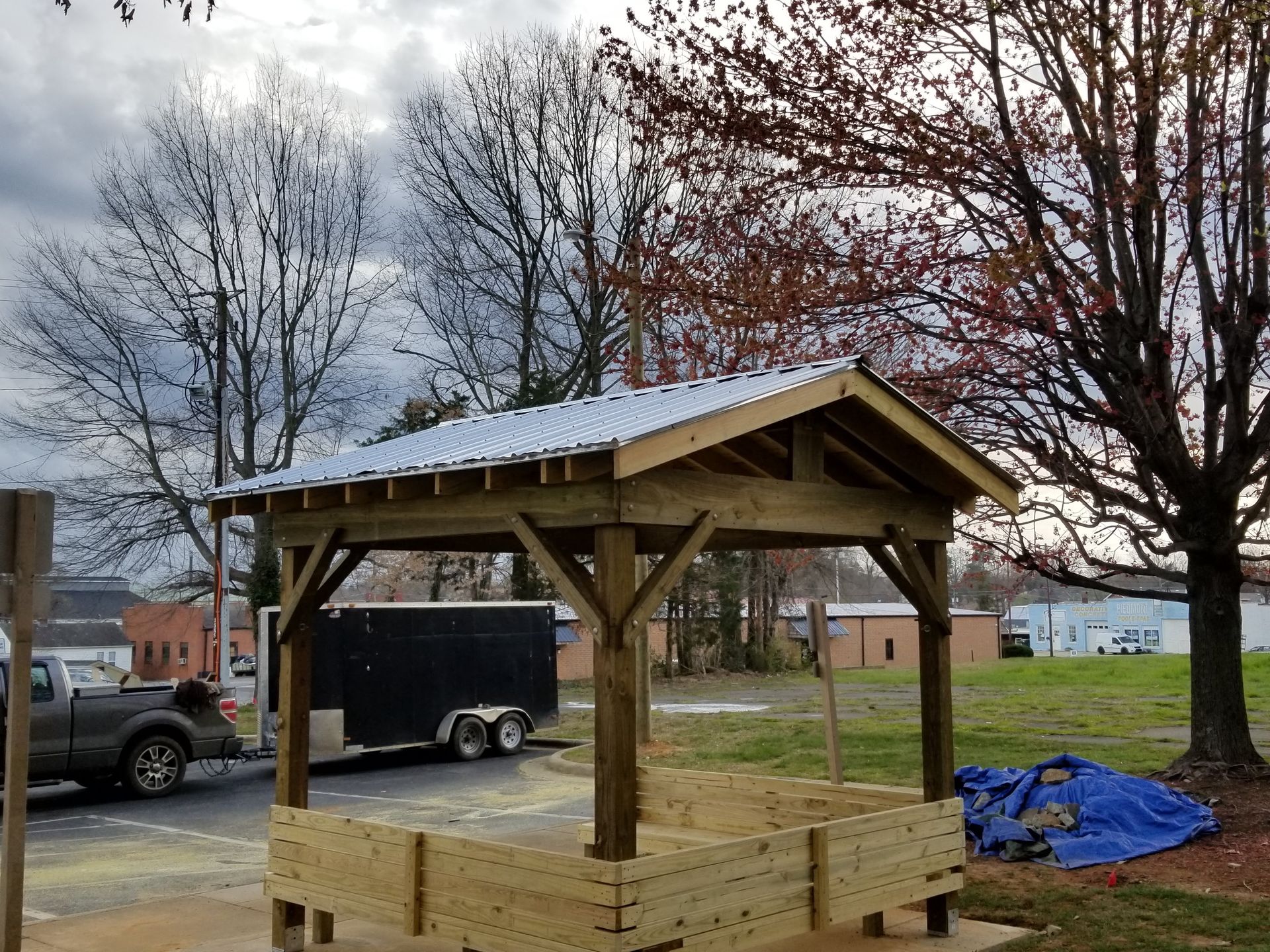 Wooden shelter with a metal roof under construction in a park. Trees and a truck are in the background.
