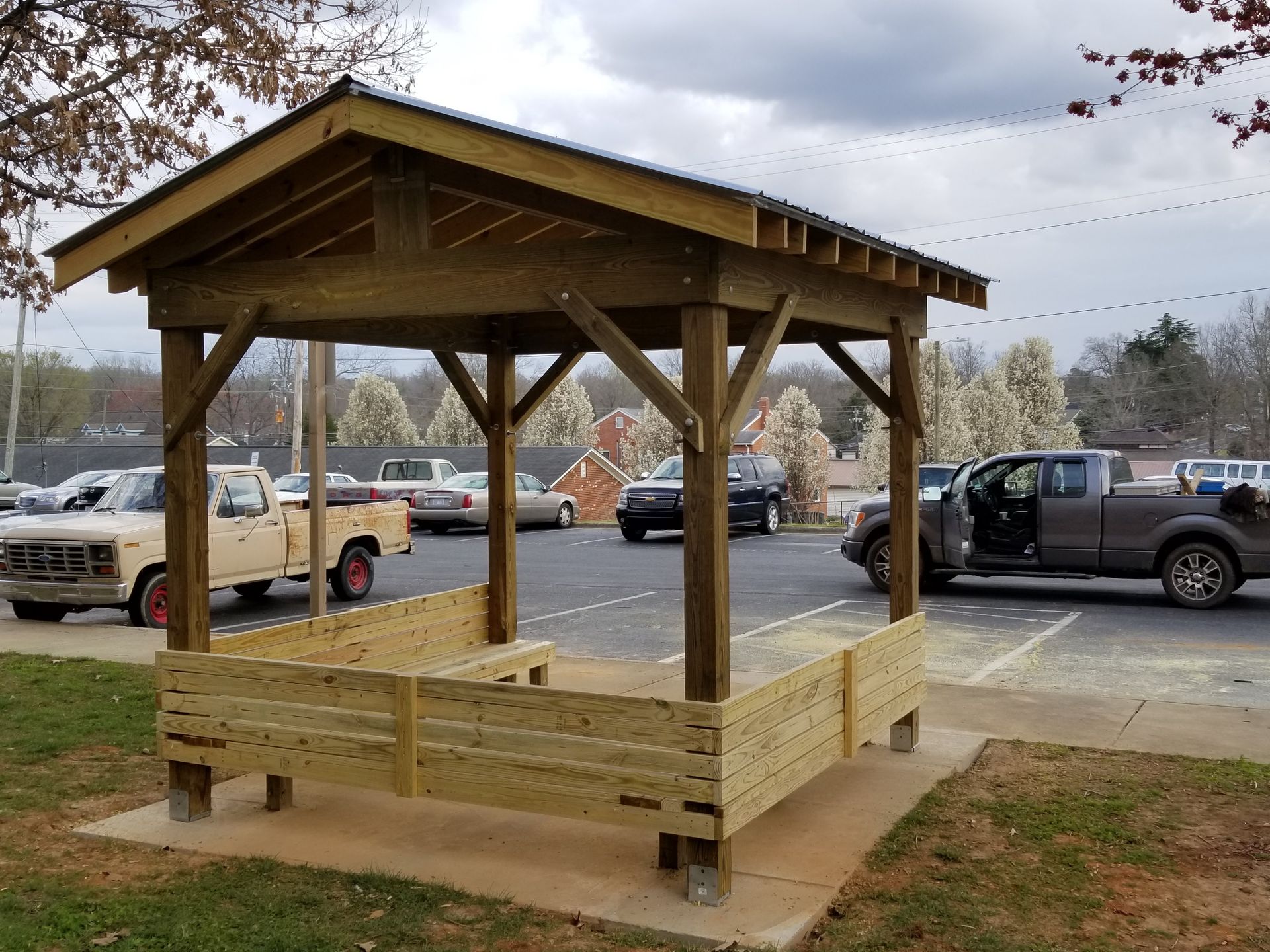 Wooden pavilion with seating in a parking lot. Brown wood, gray sky, parked cars in the background.