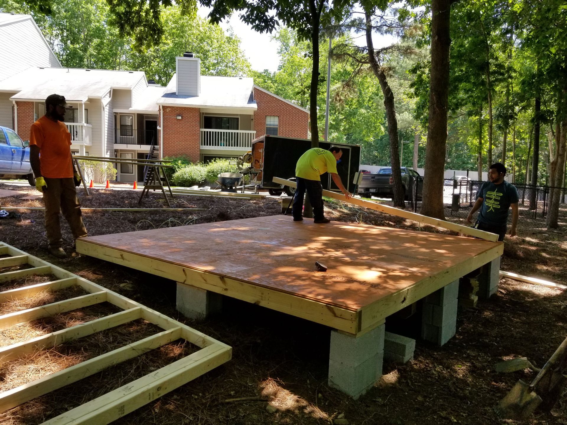 Construction workers building a wooden platform on concrete blocks in a wooded area near a building.