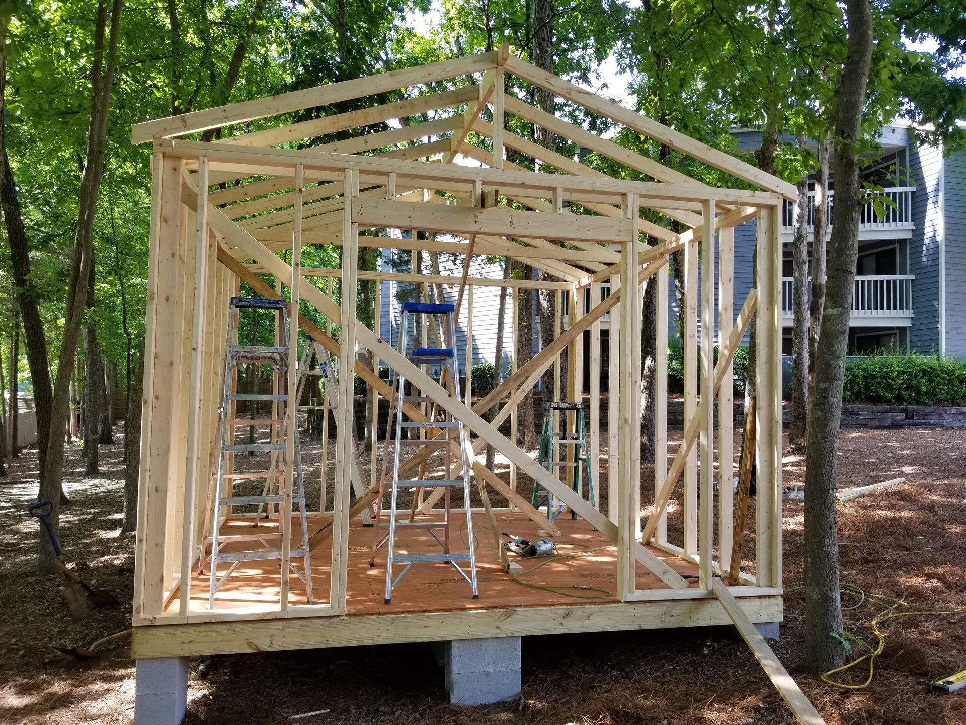 Wooden shed under construction in a wooded area with trees and a building in the background.