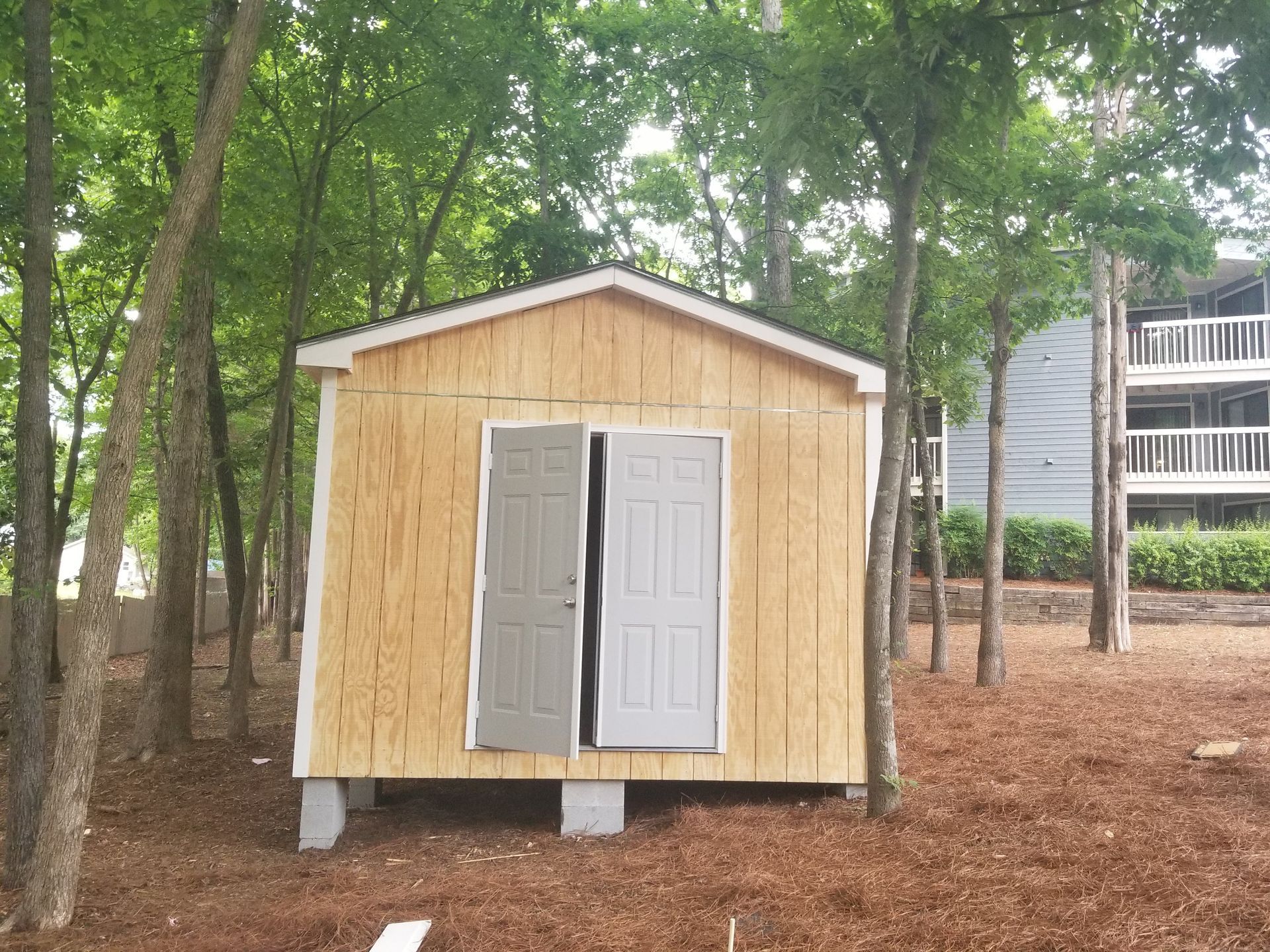 Small wooden shed with two gray doors open, set among trees and wood chips.