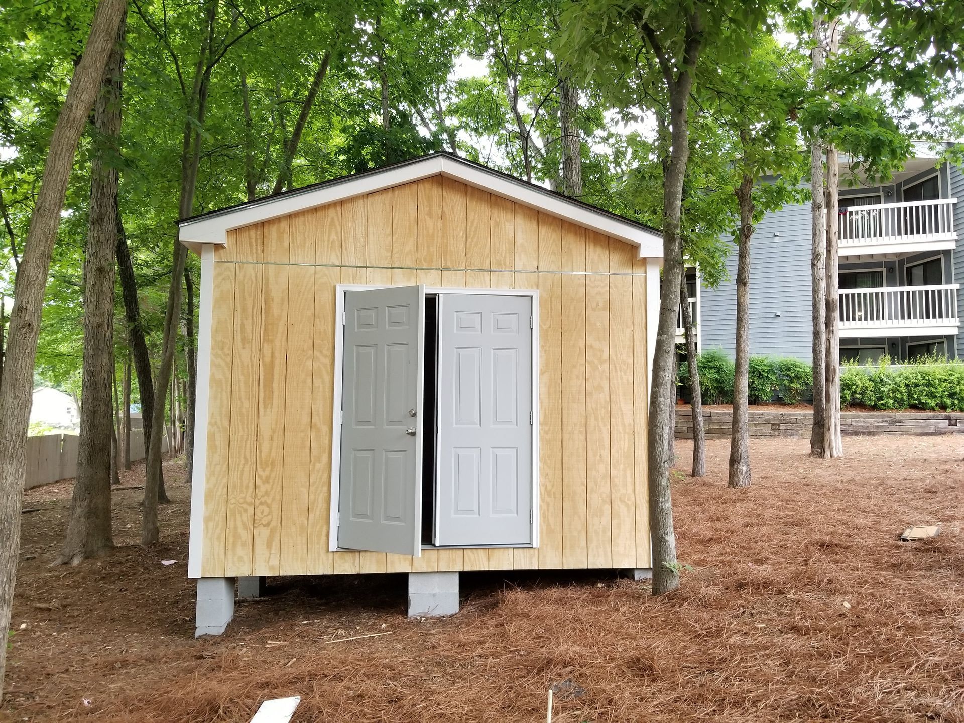 Small wooden shed with light doors, set among trees and wood chips.