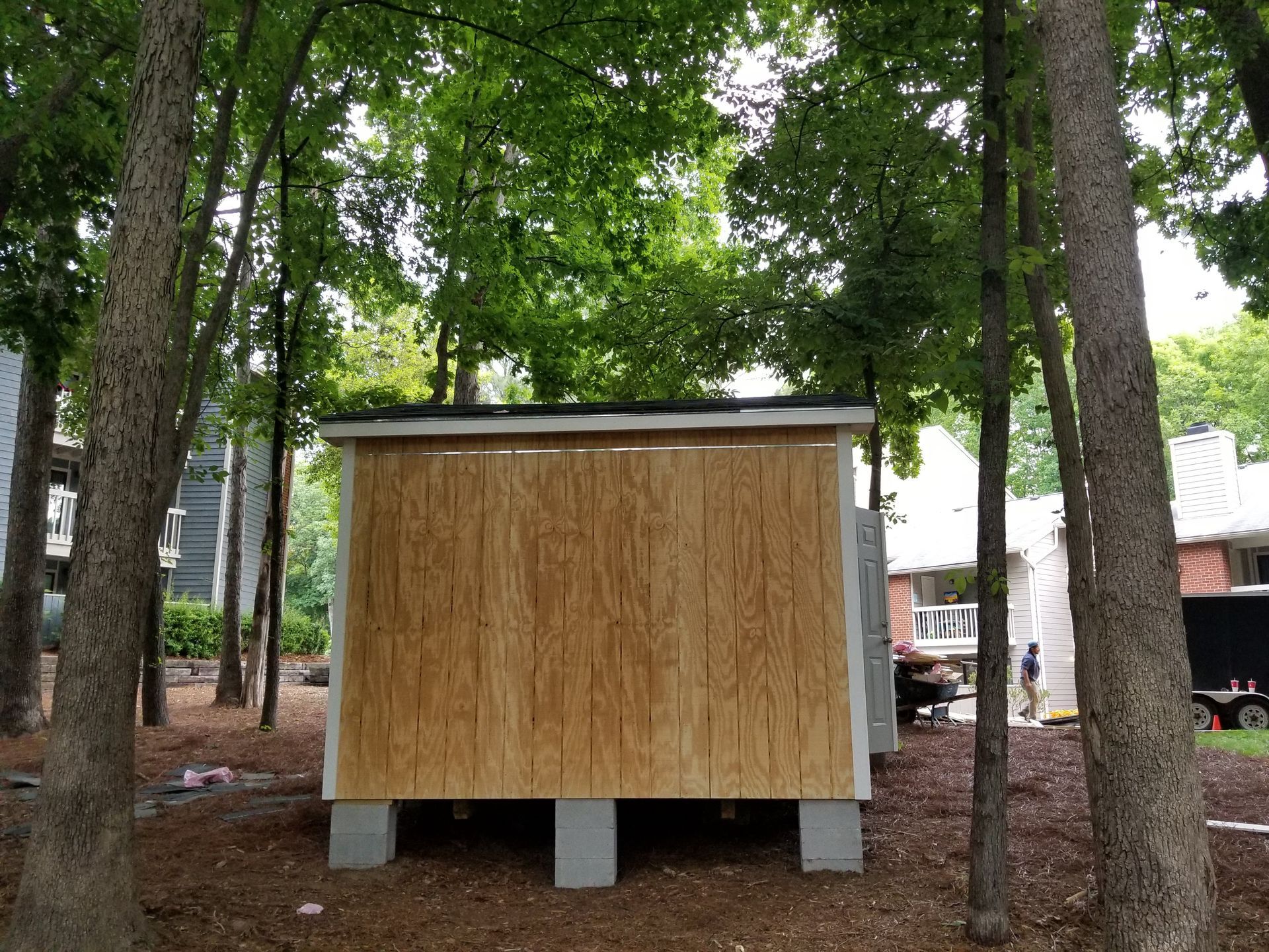 Wooden shed on concrete blocks in a wooded area with trees and a building in the background.