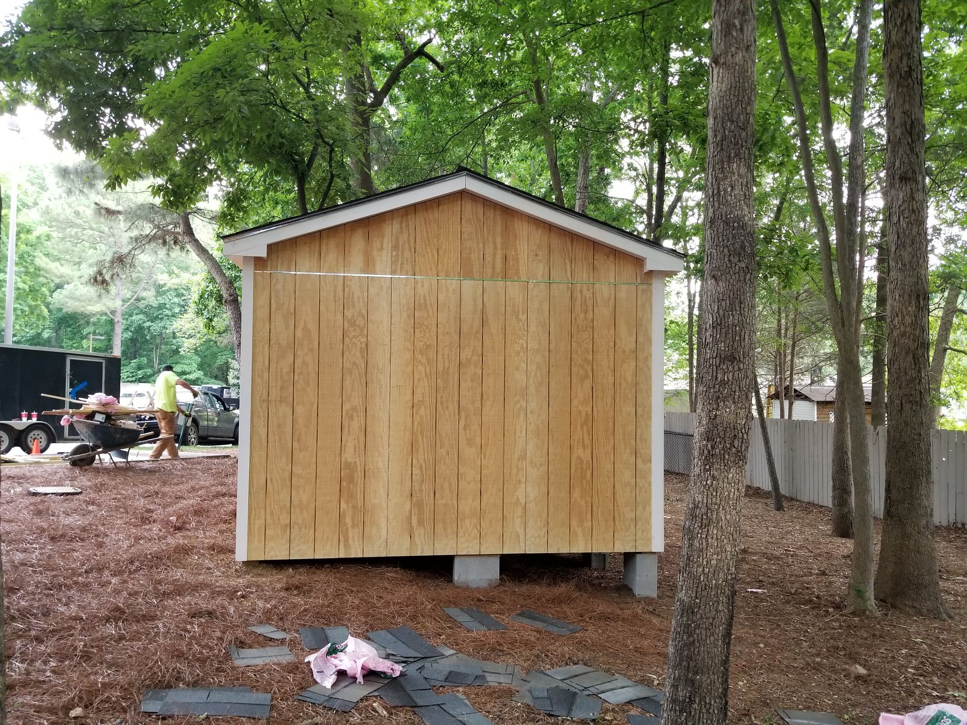 Wooden shed under construction, brown siding, white trim. Person in the background. Surrounded by trees and mulch.