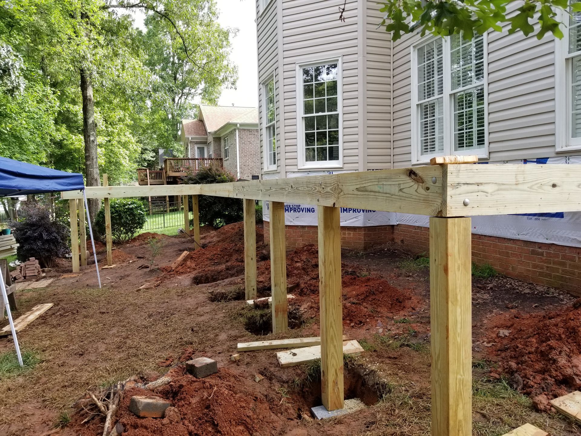 Deck construction: Wooden posts supporting a frame along a house with surrounding dirt and trees.