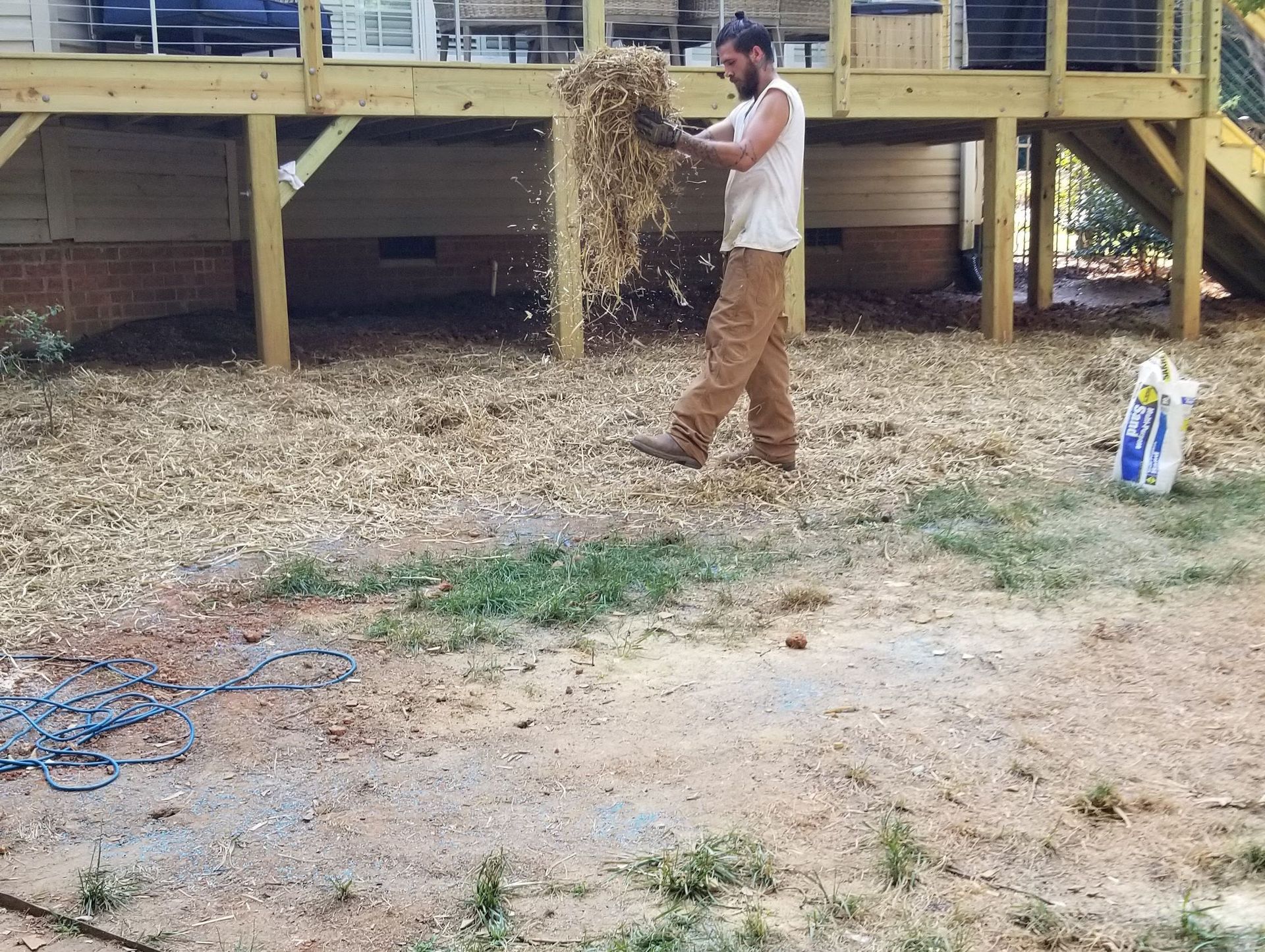 Man spreading wood chips near a deck, ground covered in chips and sparse grass, bag in background.