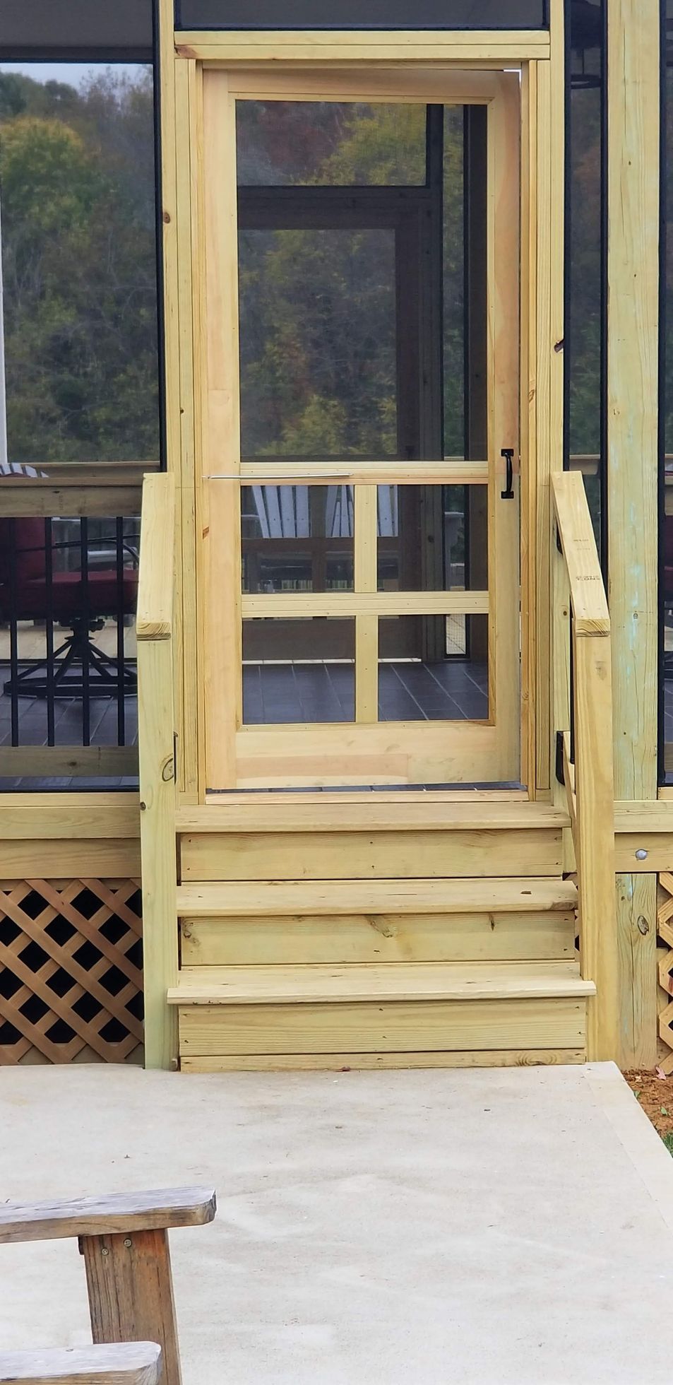 Wooden porch with steps leading to a screen door. Concrete patio with wooden bench in the foreground.
