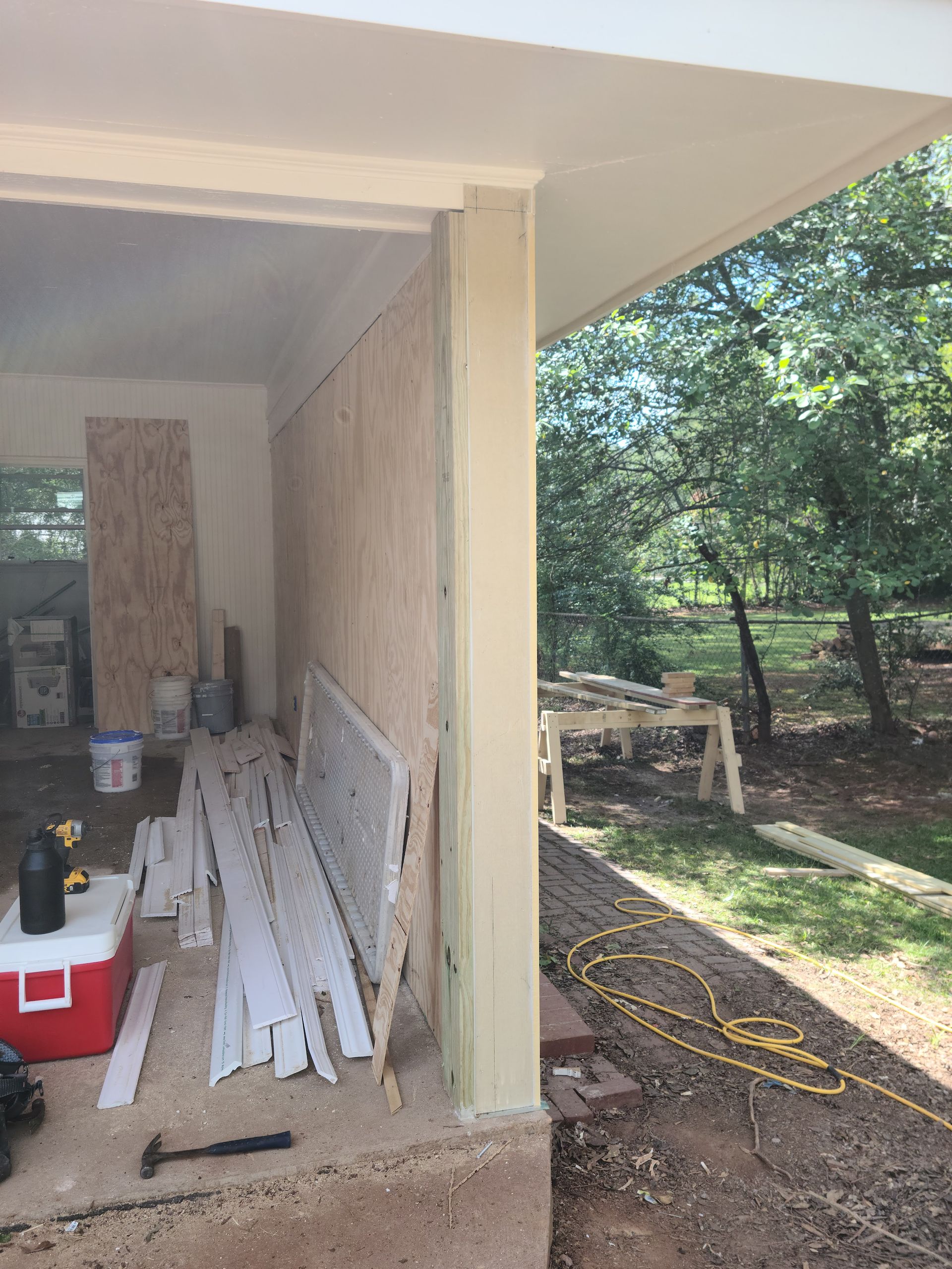 Porch under construction with exposed plywood walls and lumber, with a yard visible.