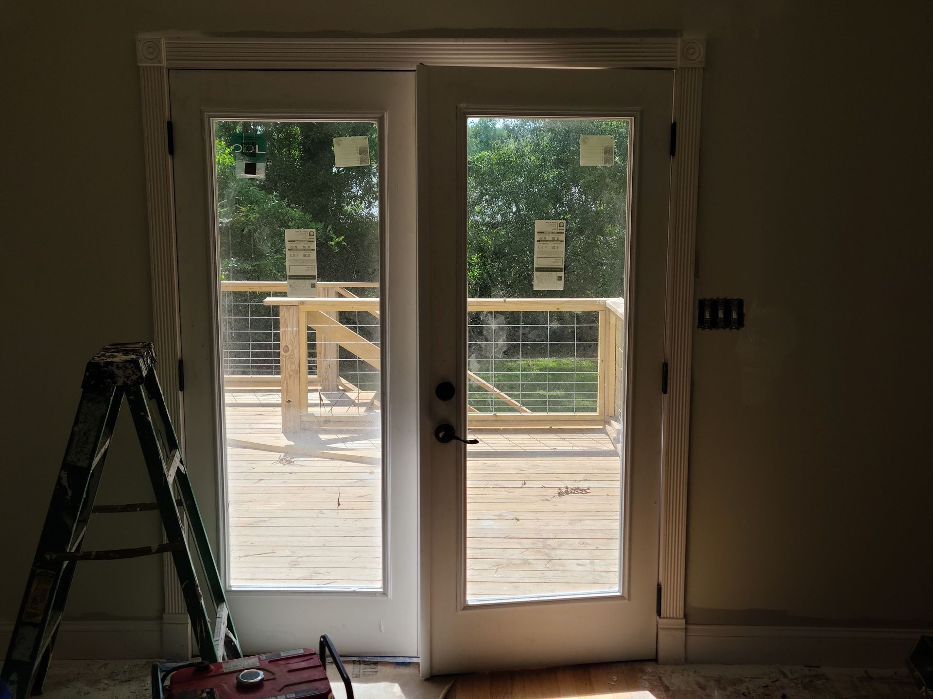 White French doors open to a wooden deck with a cable railing. A ladder leans against the left side.