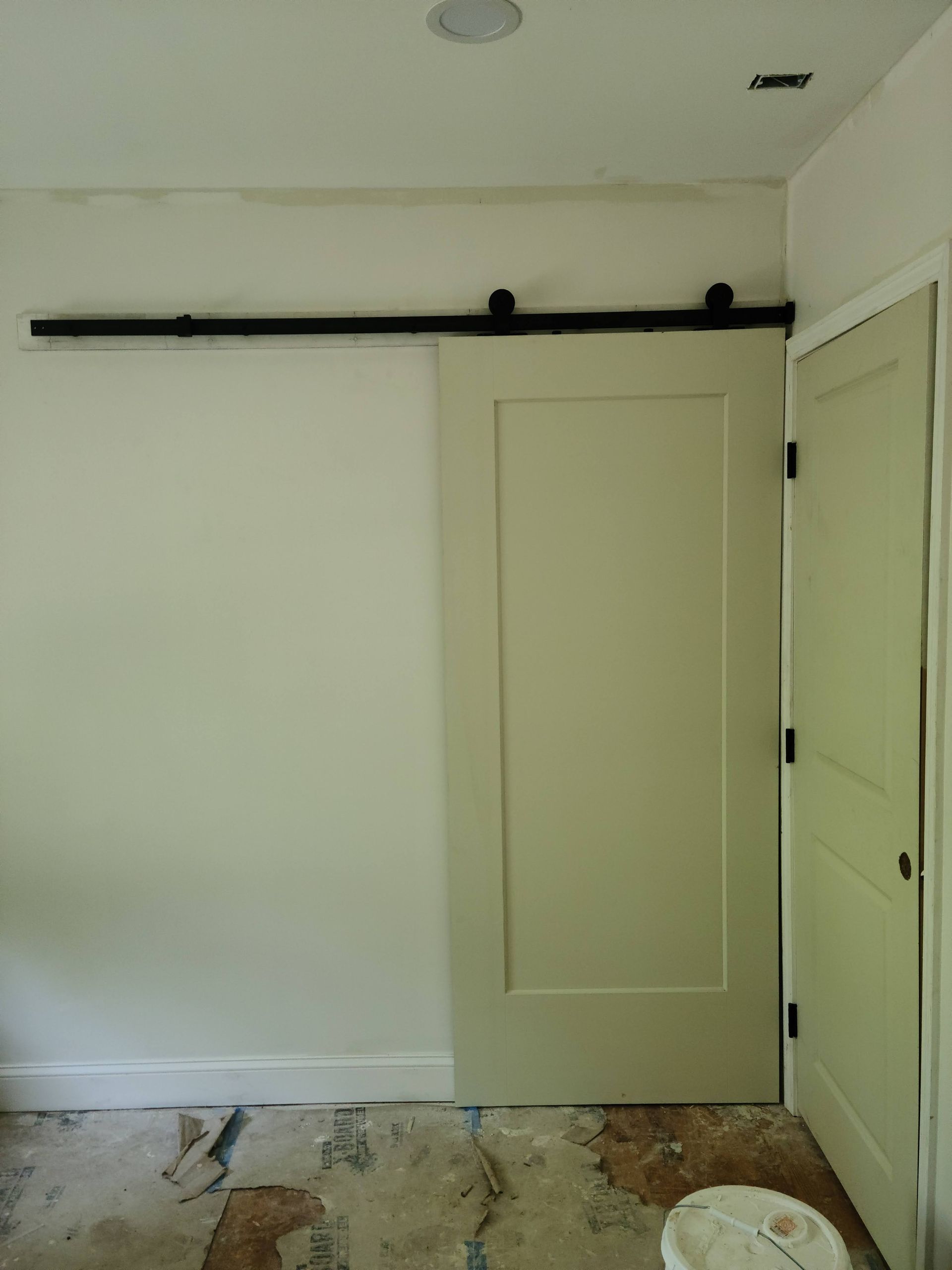 Cream-colored sliding barn door on a black track, installed in a room with a regular door and bare floor.