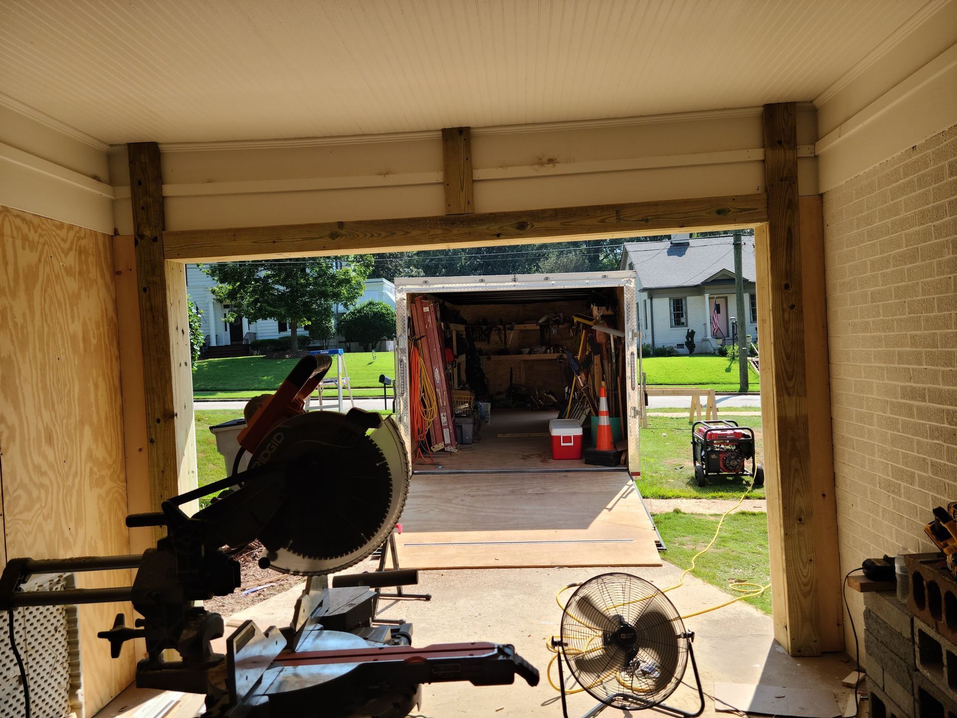 A miter saw inside a garage opening, looking out to a cluttered garage, green lawn, and houses.