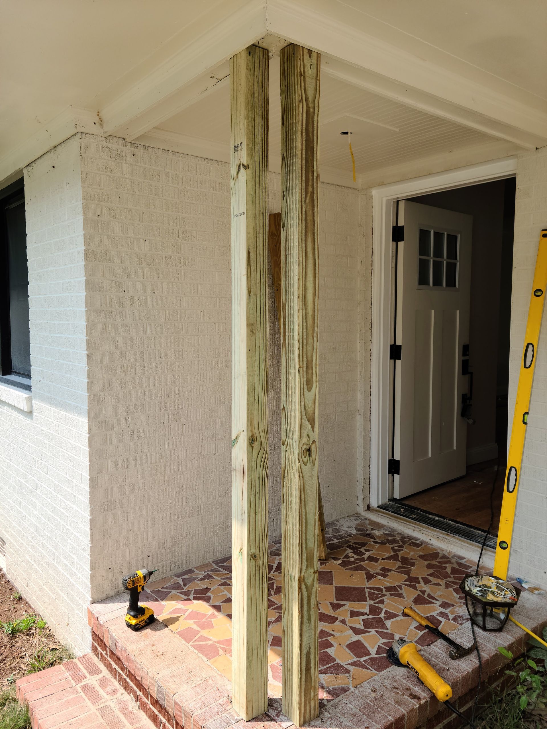 Construction of a porch column, using wooden posts, next to a textured white wall and doorway. A level leans against the wall.