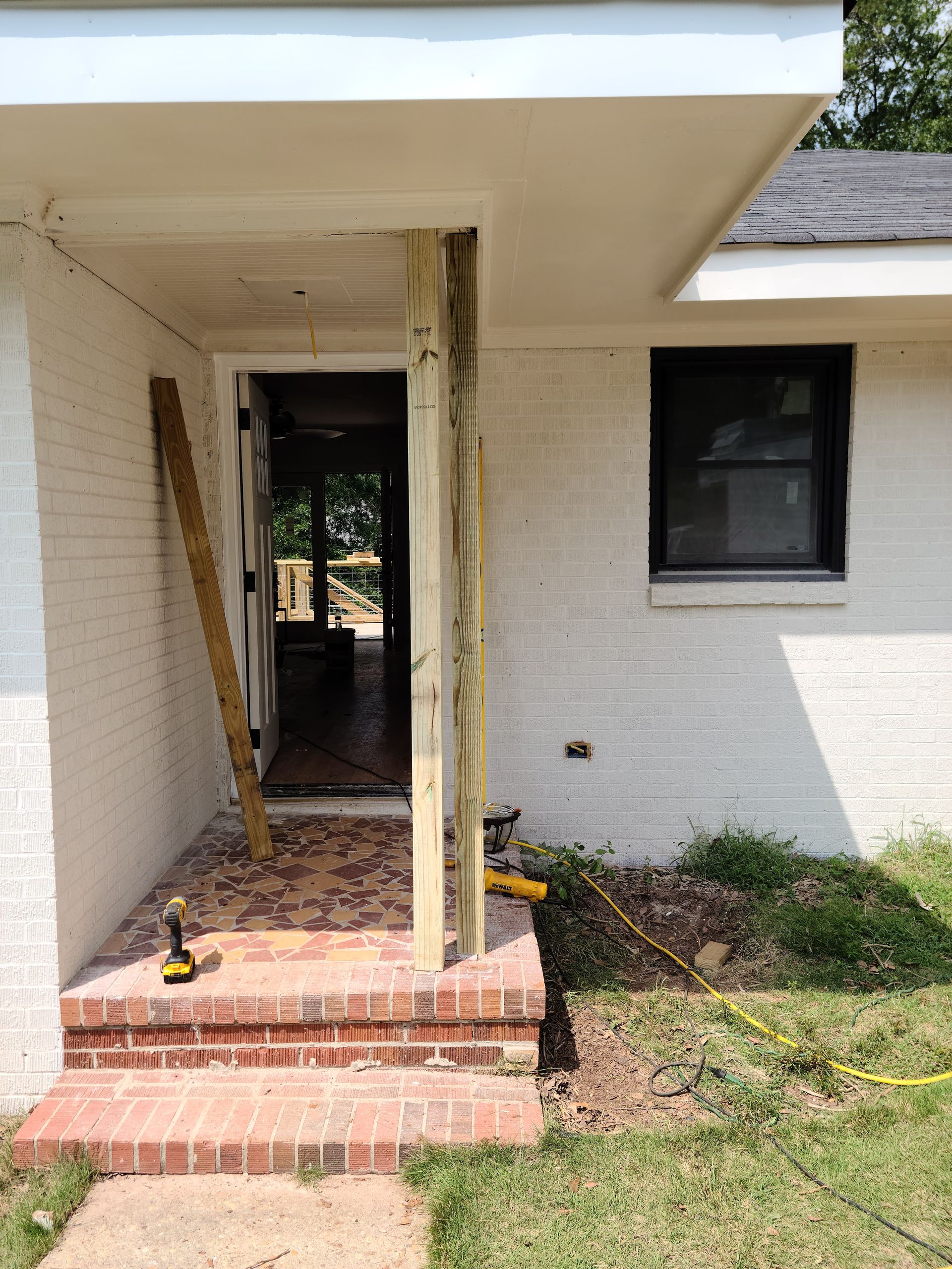 Exterior view of a home's entryway under construction; brick steps, a support beam, and tools visible.