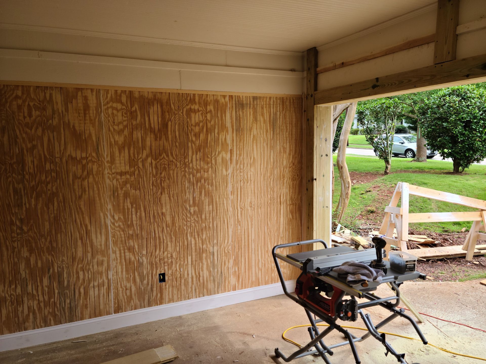 Interior of a garage under construction with plywood walls, a table saw, and an open doorway.