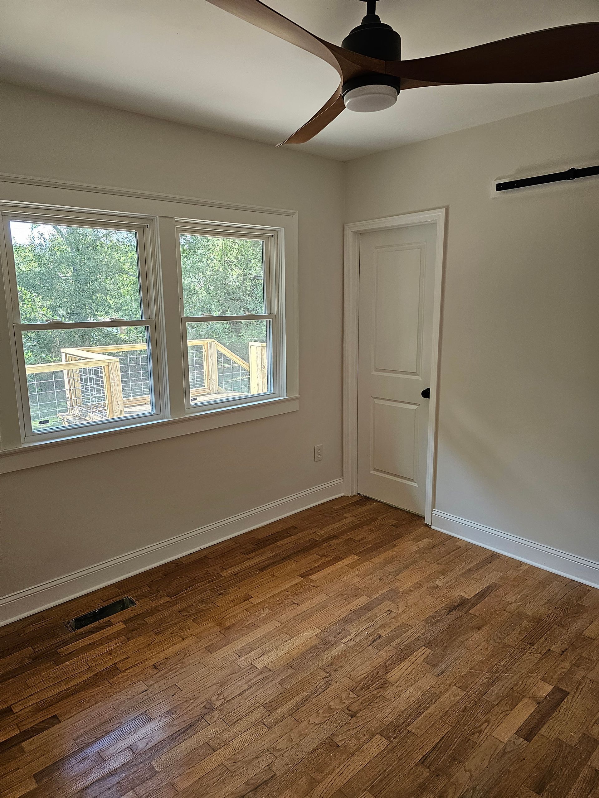 Empty bedroom with wood floors, window, white door, and ceiling fan.