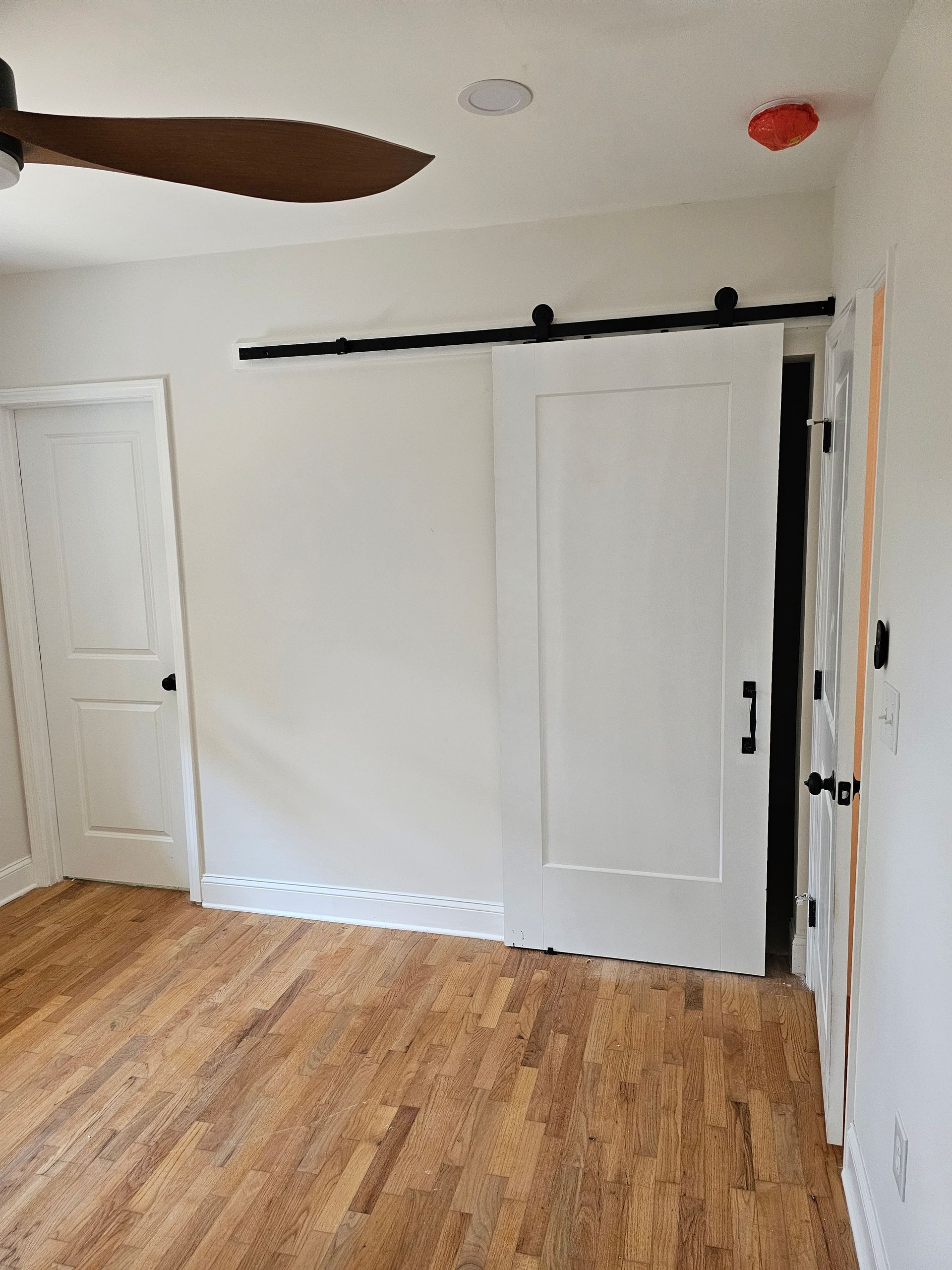 Bedroom with hardwood floors, white walls, and a sliding barn door.  A closed door is on the left, open door on the right.