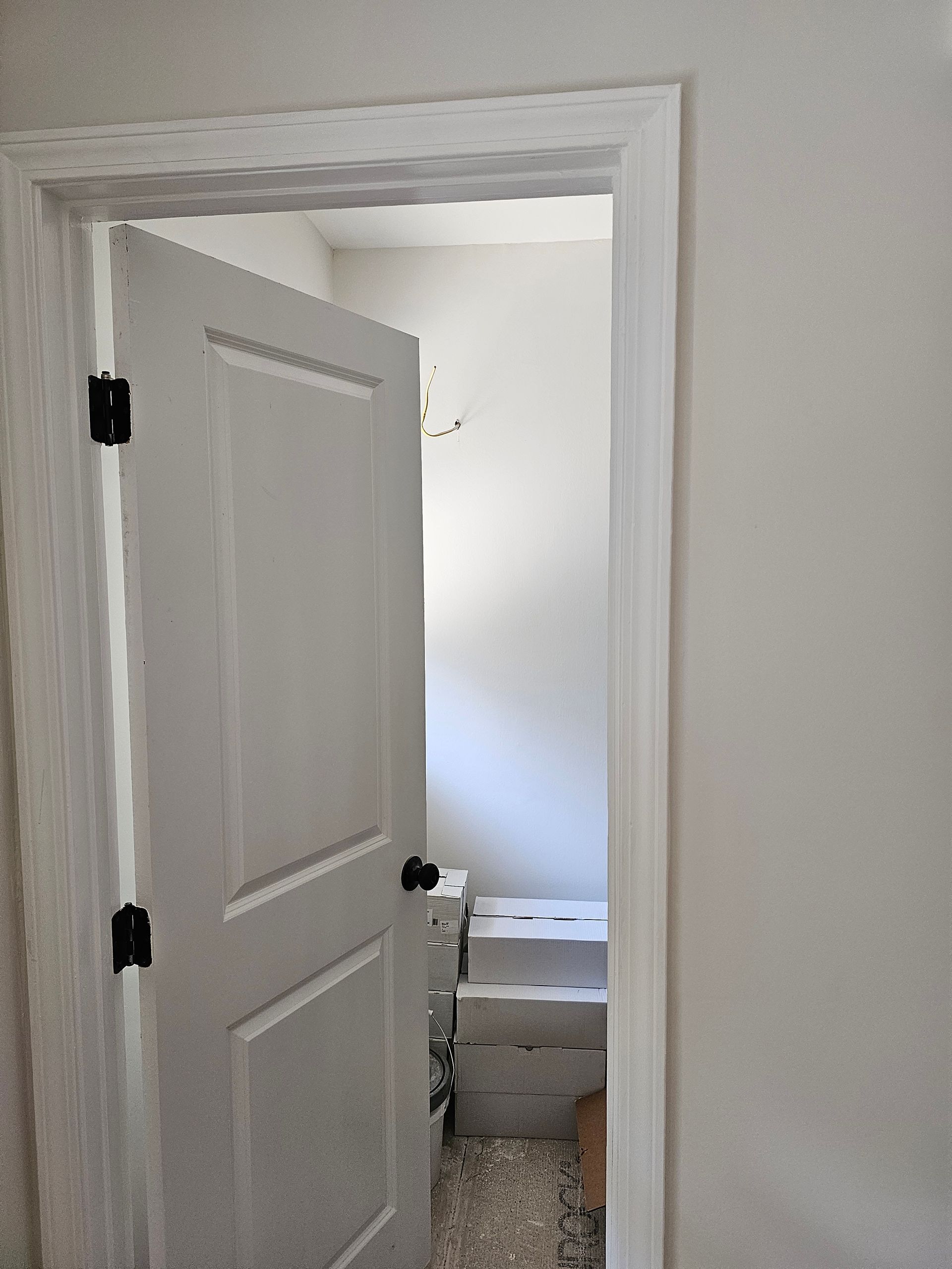 White door, ajar, with black hinges and knob, leading to a small, cluttered white-walled room with boxes.