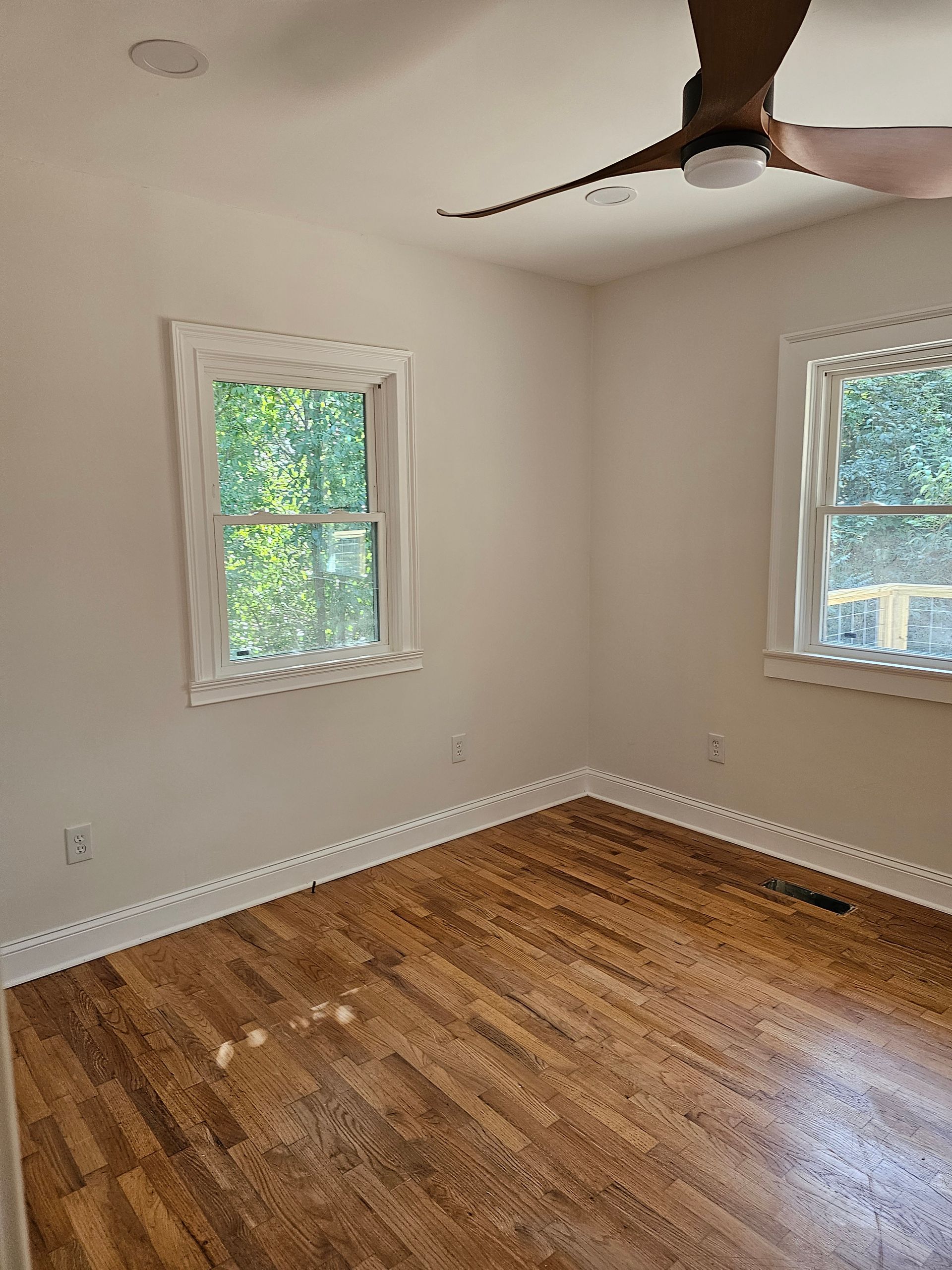 Empty bedroom with hardwood floors, two windows with white trim, and a ceiling fan.