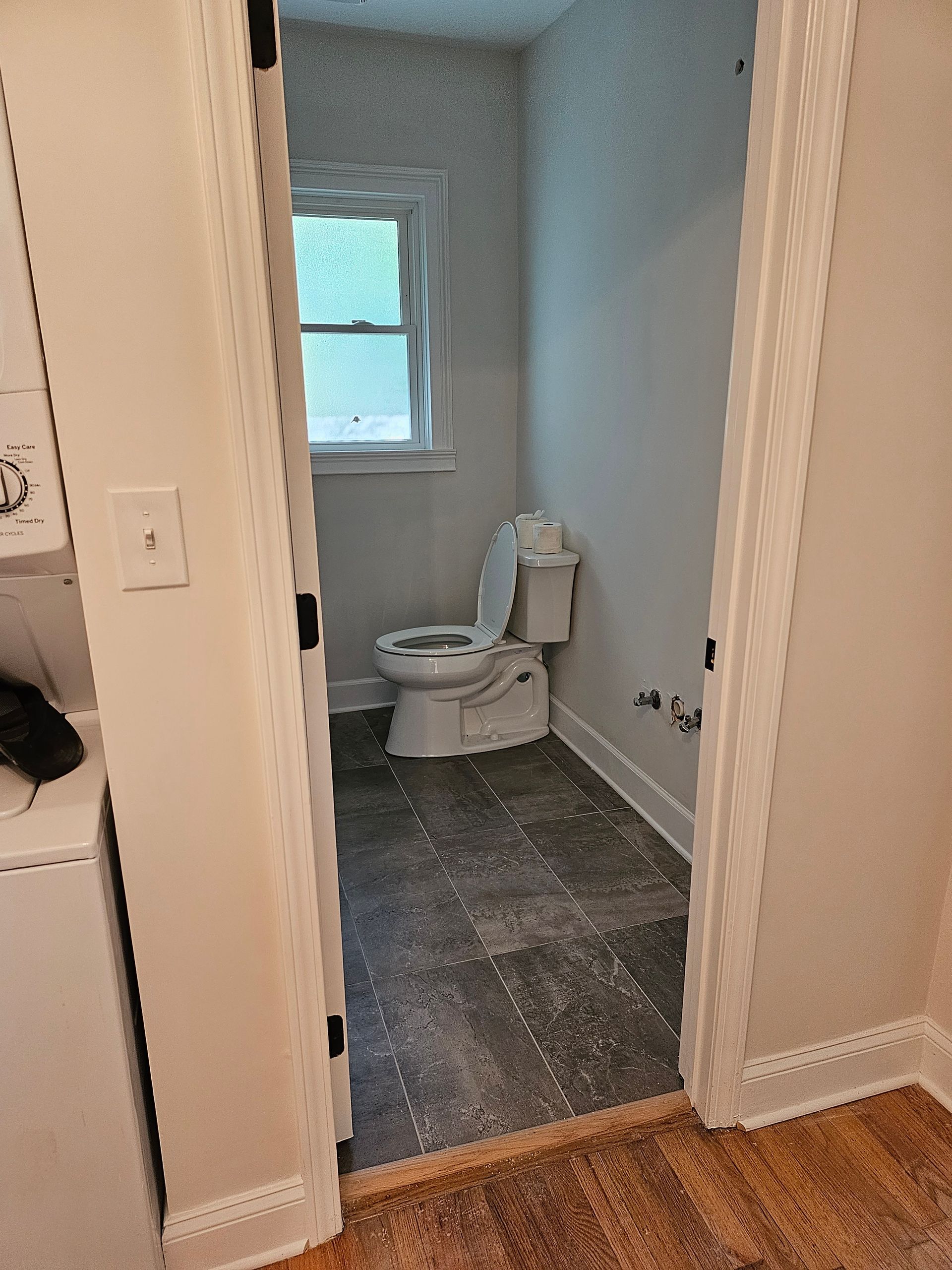 Small bathroom with open door, toilet, window, and gray flooring.