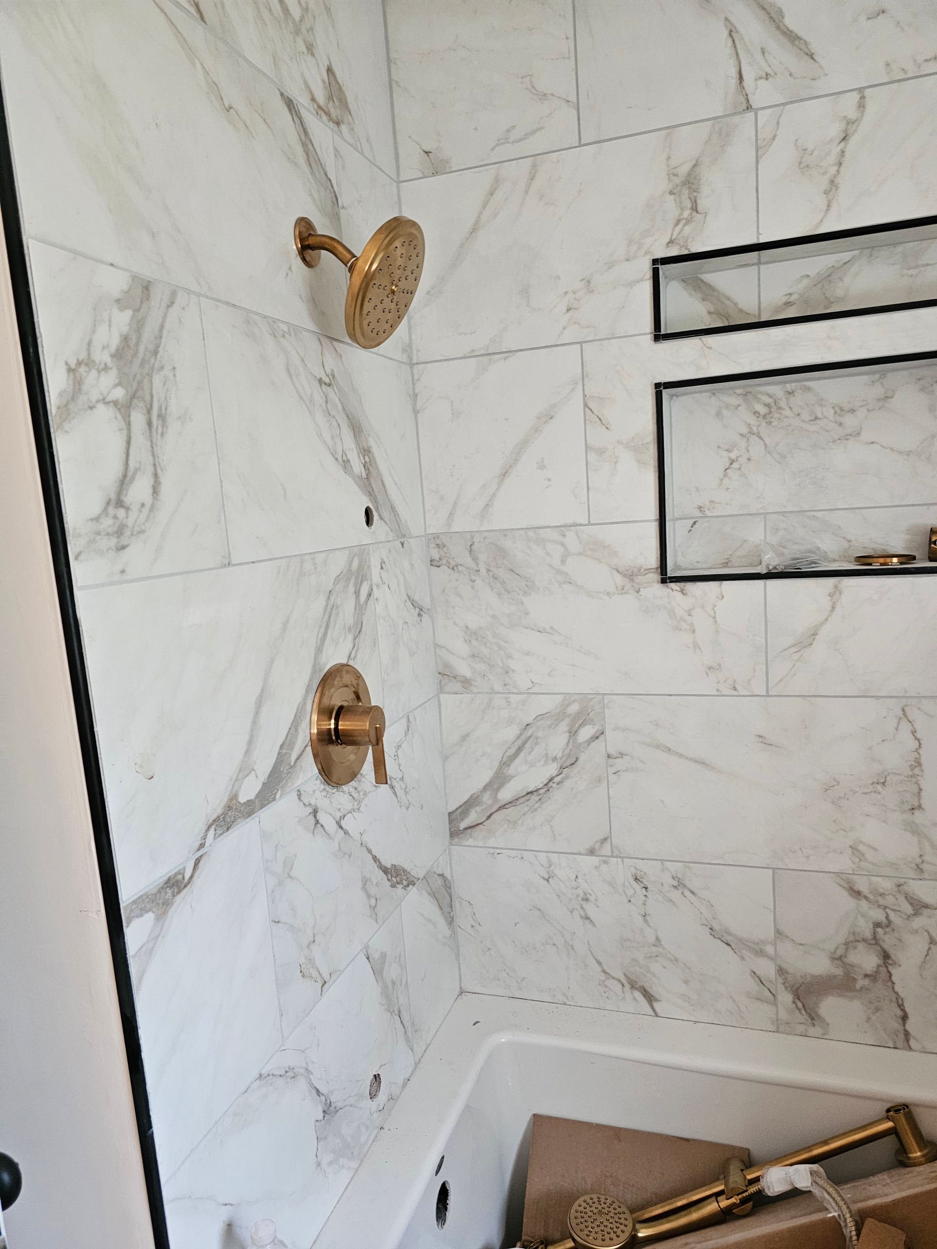 Gold shower fixtures on white marble tile walls in a bathroom. Two built-in shelves.