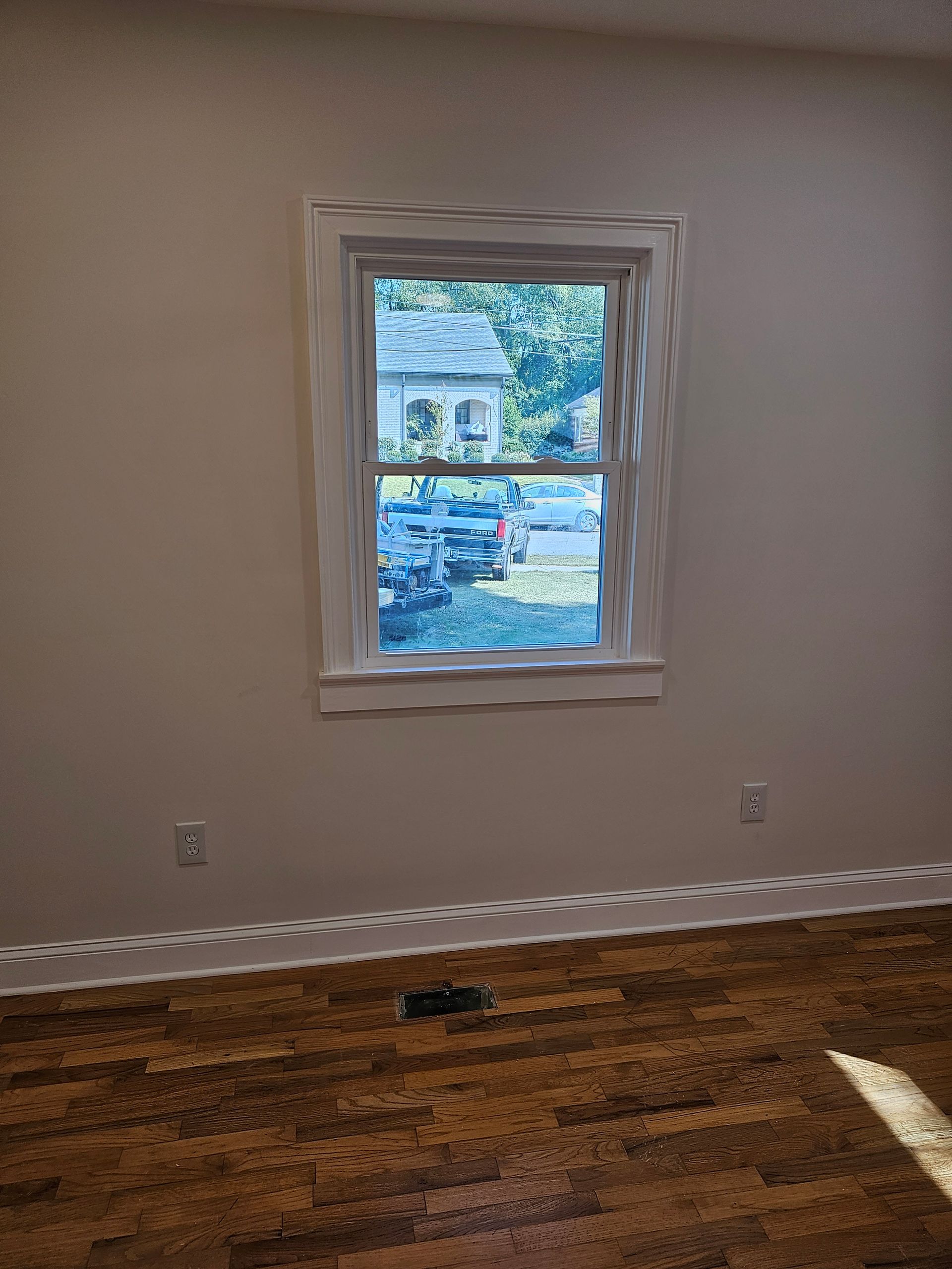 Window in a beige-walled room with a view of a building and greenery; brown wood floor.