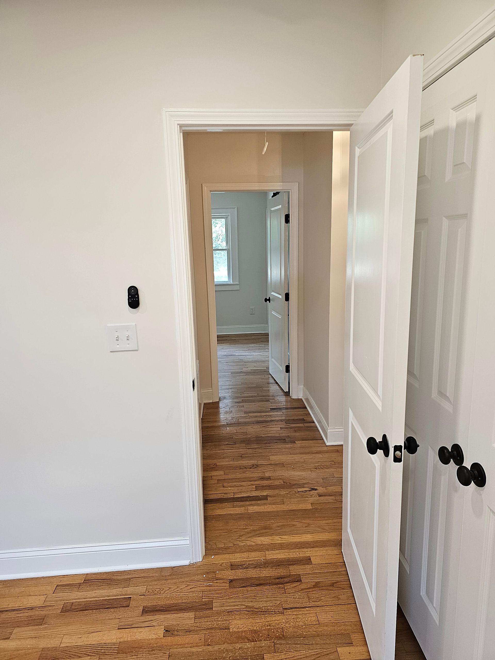 Hallway with open doors; white walls, brown flooring, and a view into another room with a window.