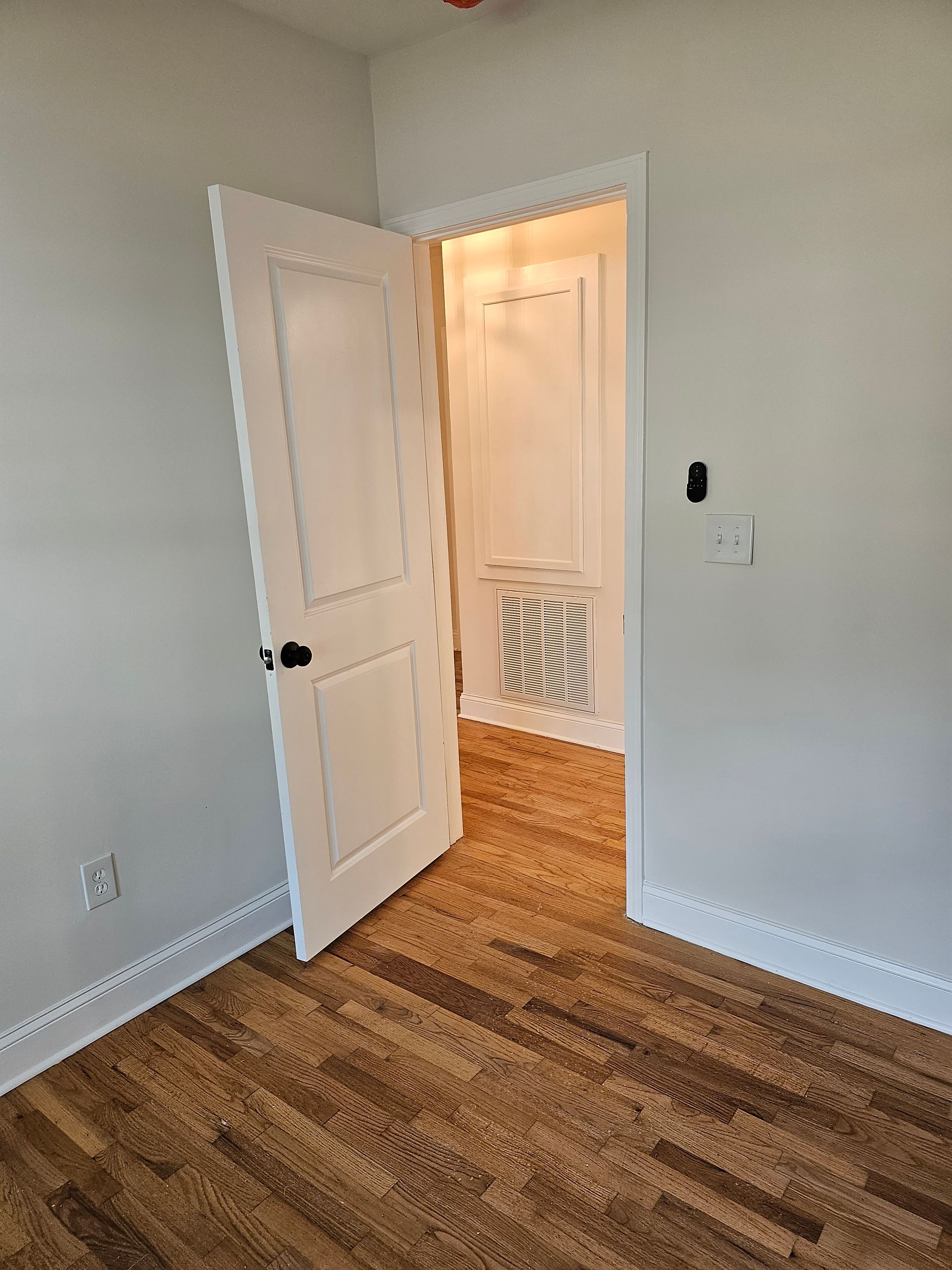 A bright, empty room with hardwood floors and a partially open white door leading to a smaller room.