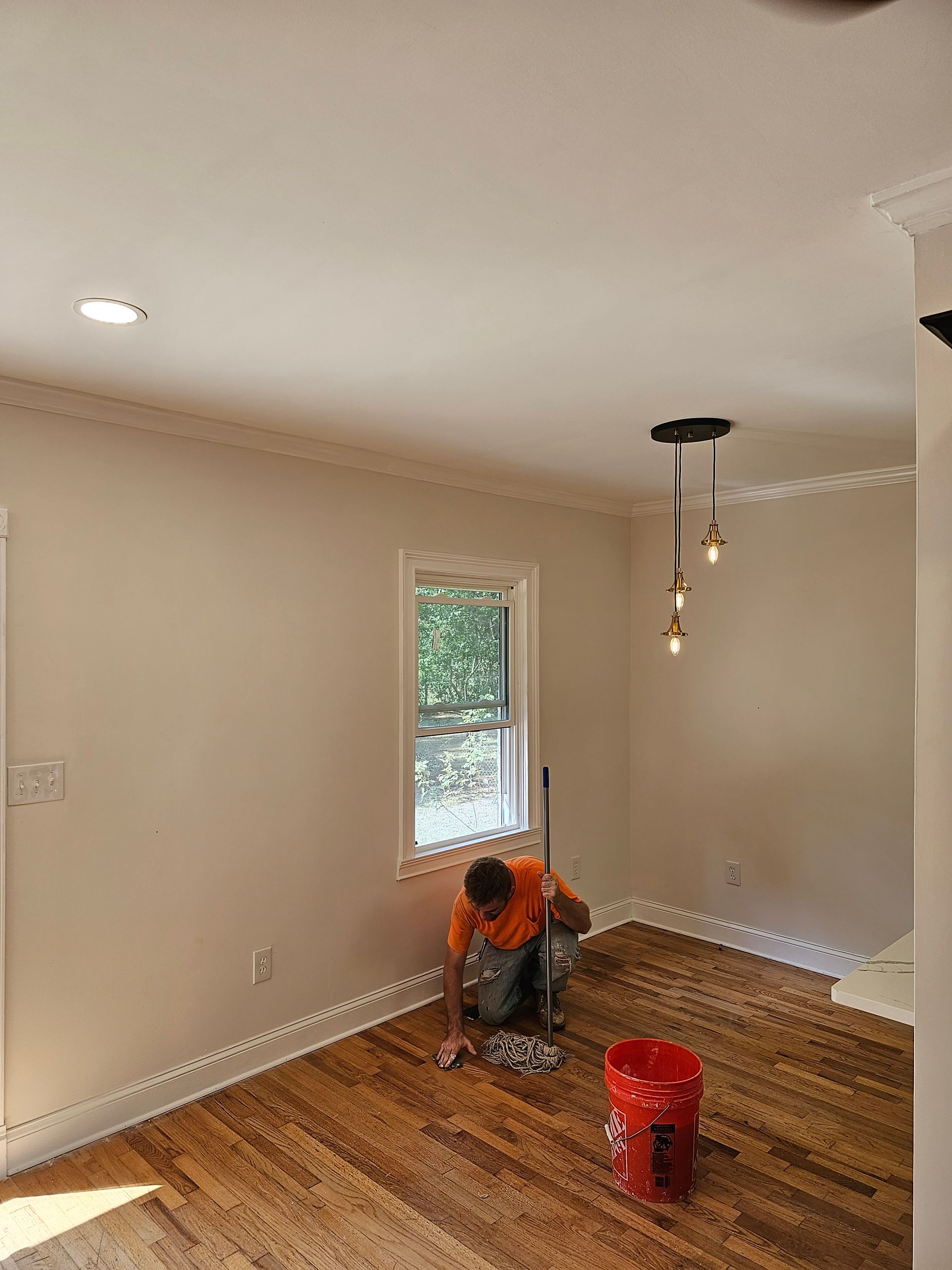 Person kneeling, wiping hardwood floor in a room with a window, ceiling lights, and red bucket.