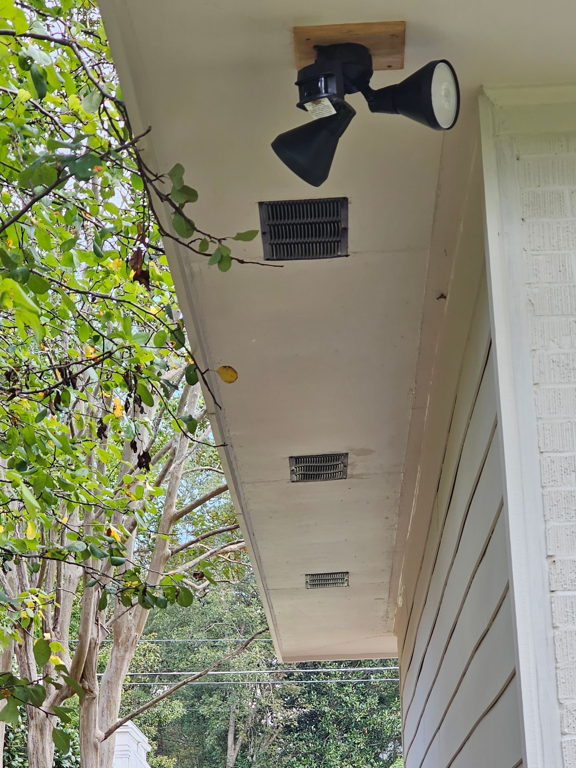 Floodlights mounted on wooden block, beneath a home's overhang, near a ventilation grate and siding.