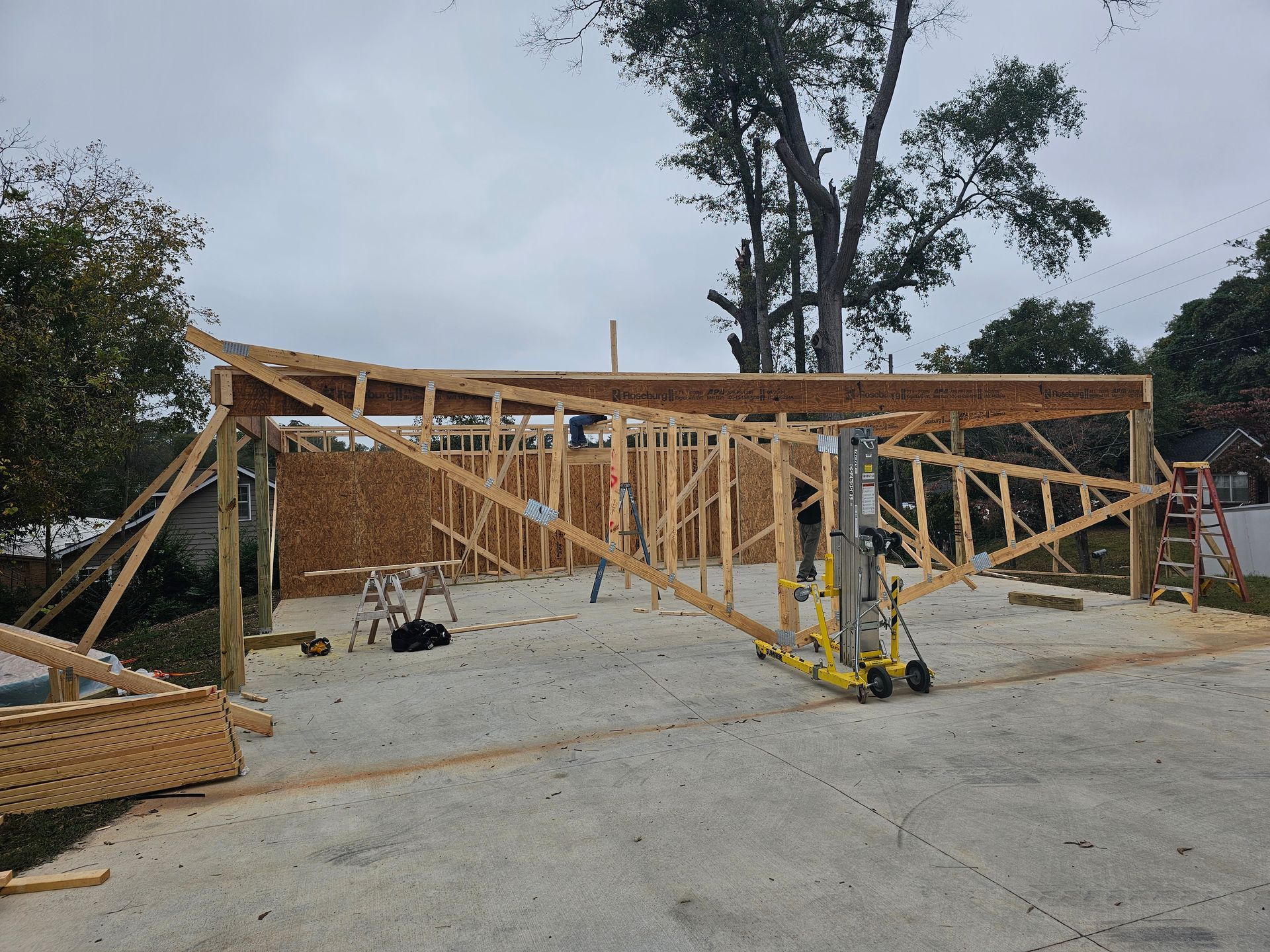 Construction of a wooden building frame outdoors. A lift, lumber, and a cloudy sky are visible.